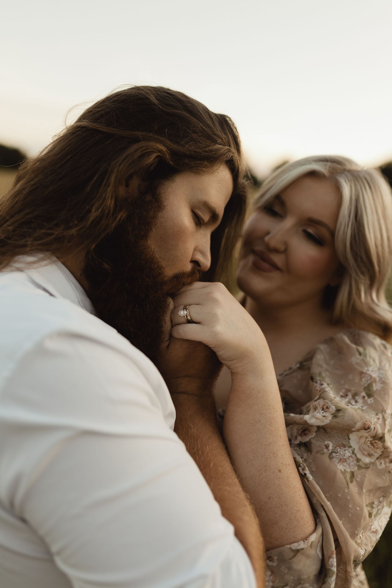 A bearded man with long hair kisses a woman's hand while she smiles warmly, wearing a floral dress outdoors.