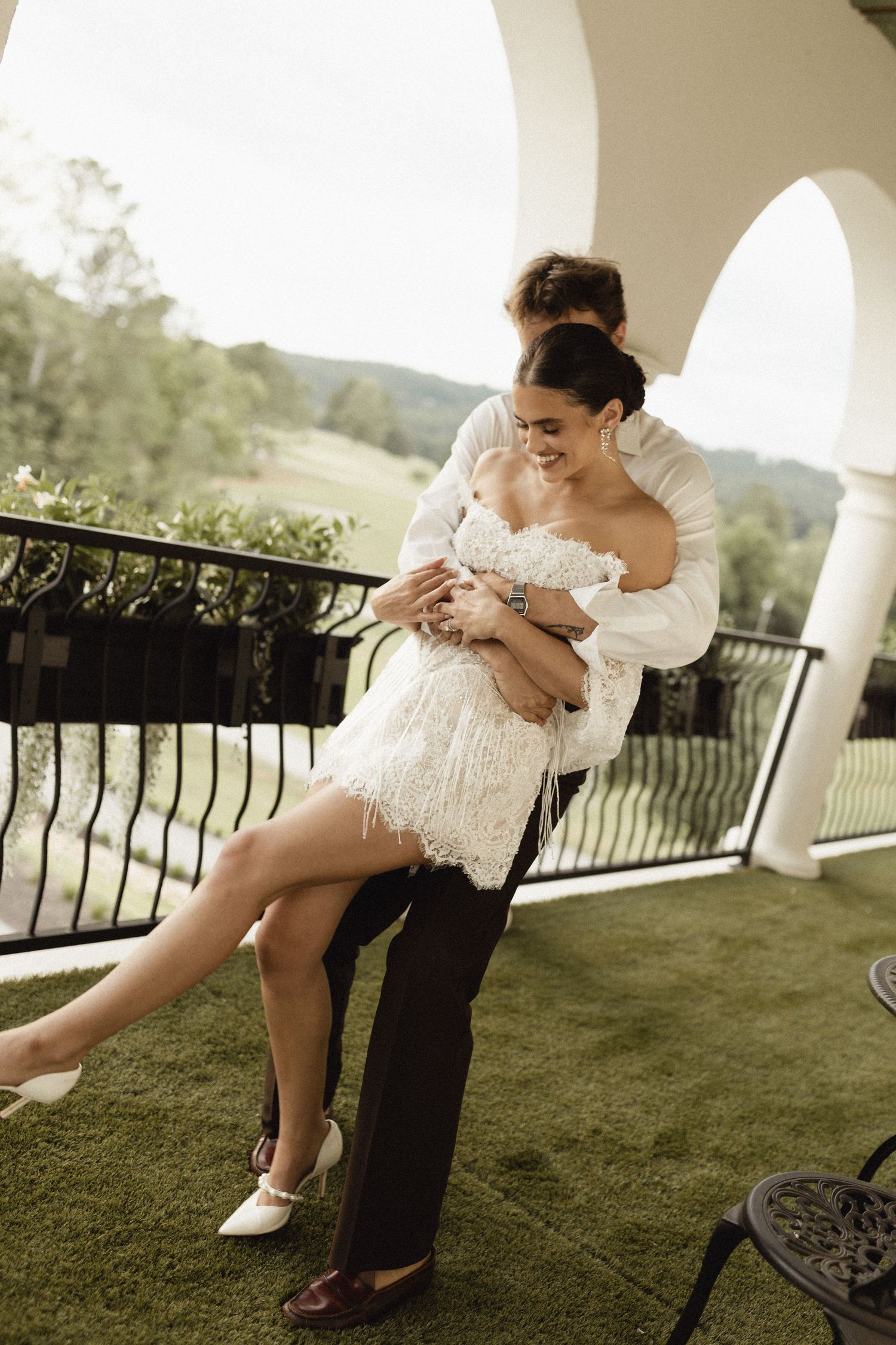 Bride in white lace mini dress being lifted by groom on elegant balcony with green landscape views.