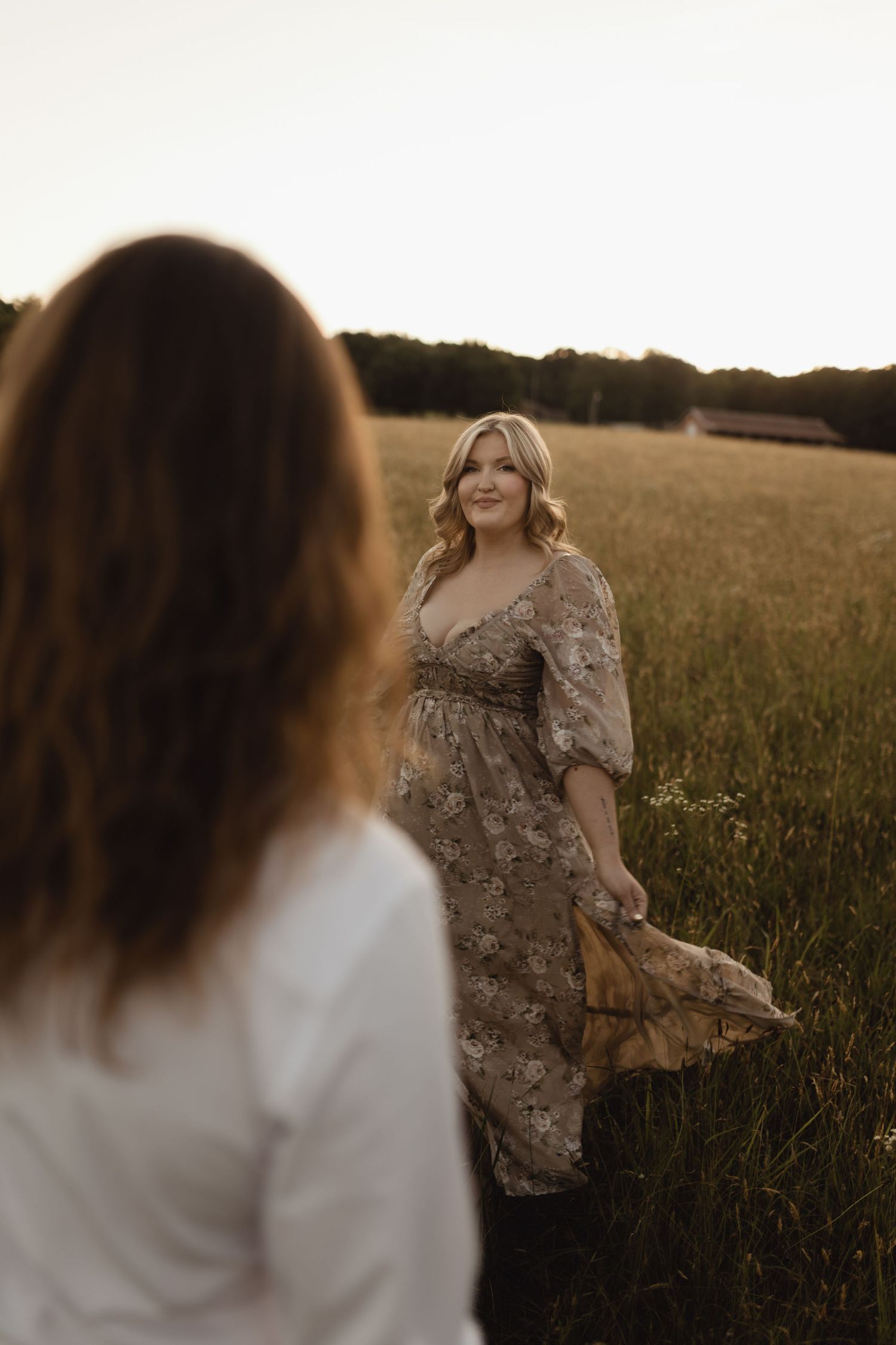 Two women in a golden field at sunset, one in a floral dress holding its hem, smiling warmly at the other.