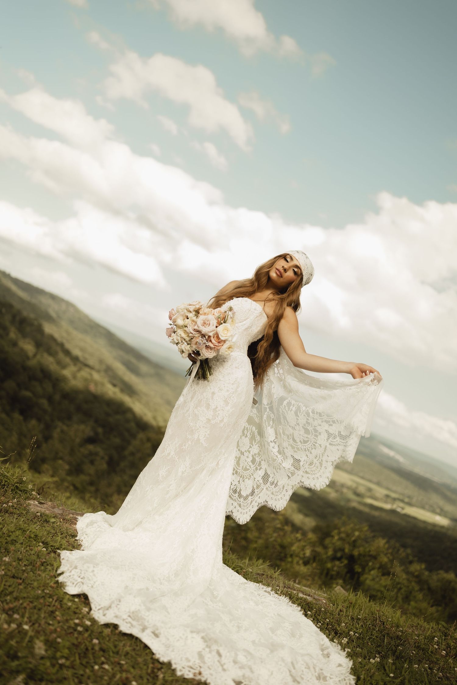 Bride in lace wedding dress holding bouquet poses on mountain hilltop with scenic valley and cloudy sky backdrop.
