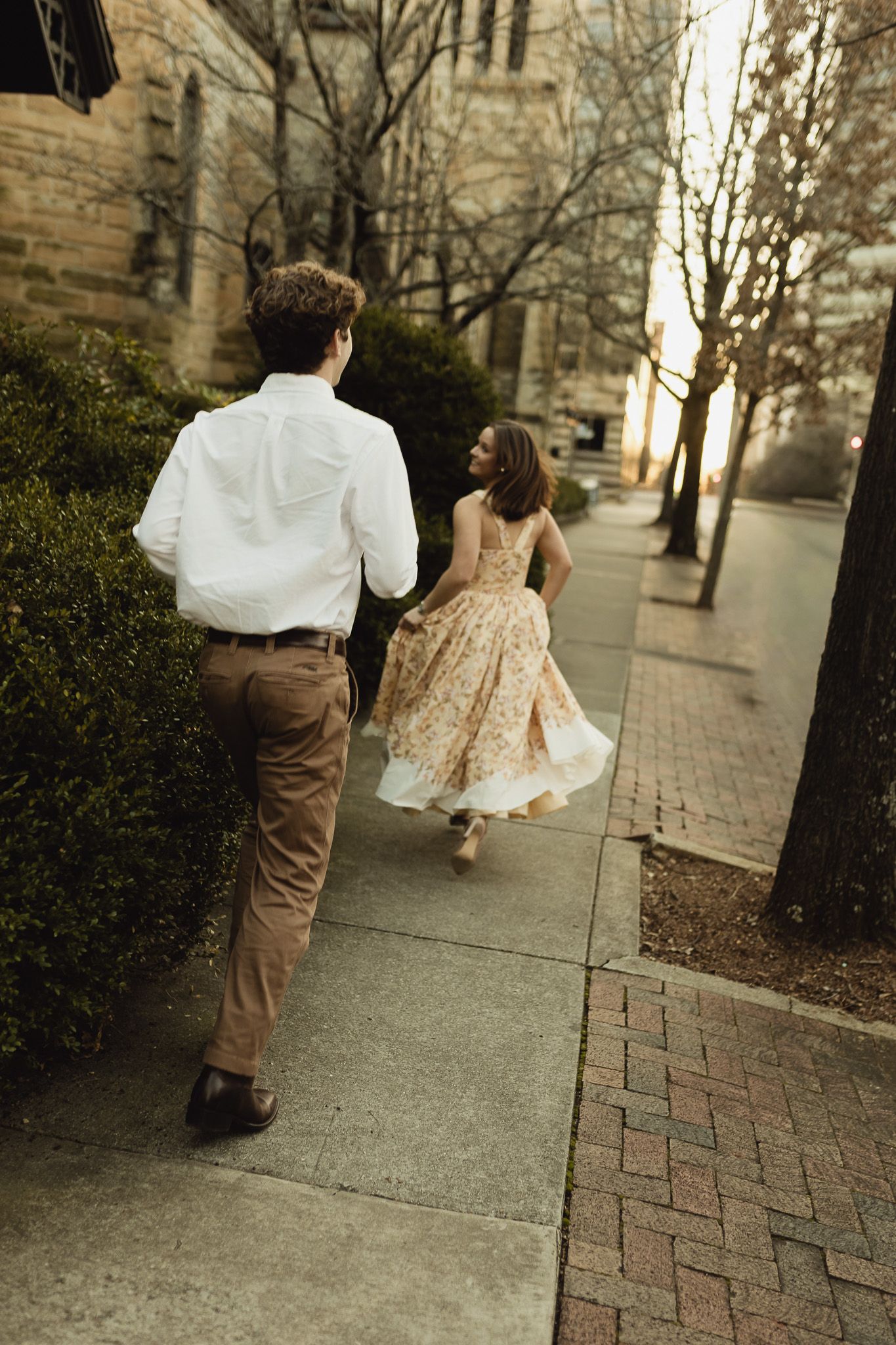 A couple runs hand-in-hand down a tree-lined sidewalk, the woman in a floral dress and the man in a white shirt.