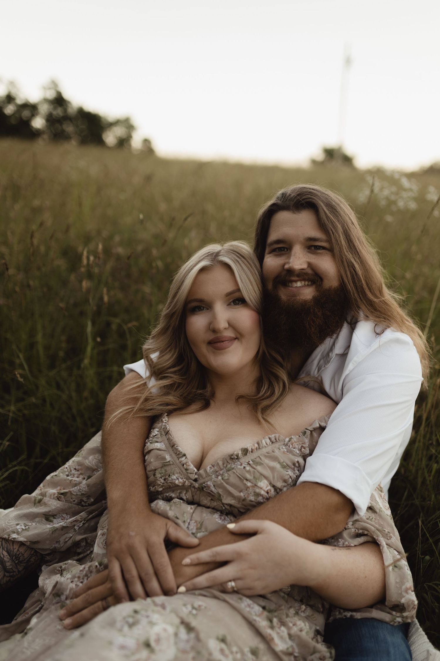 A couple sits in a lush green field, woman in floral dress, bearded man with long hair embracing her warmly.