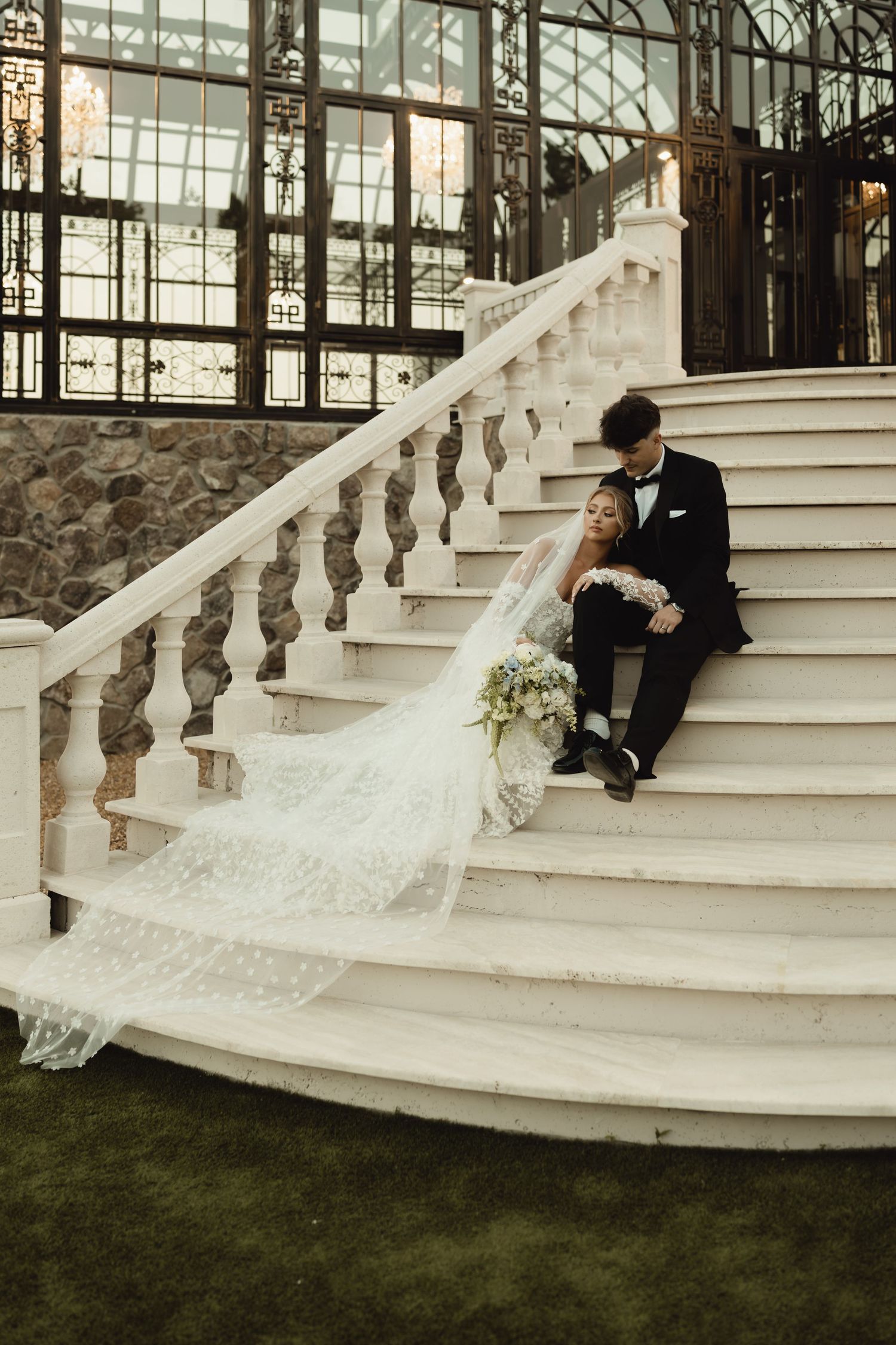 Bride and groom sitting on elegant white marble staircase, bride's lace veil flowing down steps, bouquet in hand.