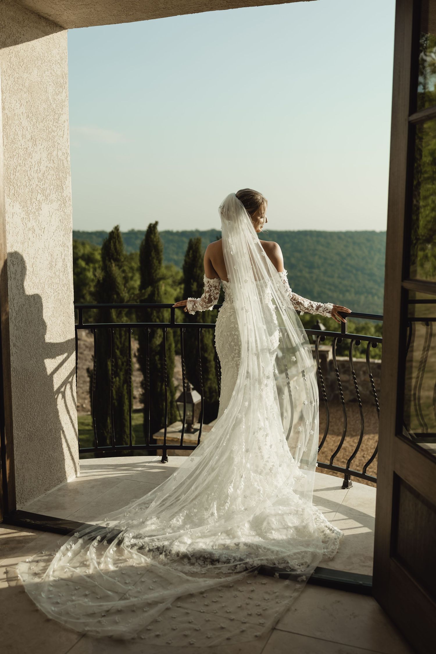 Bride in lace wedding gown and long veil stands on balcony overlooking lush green landscape on sunny day.