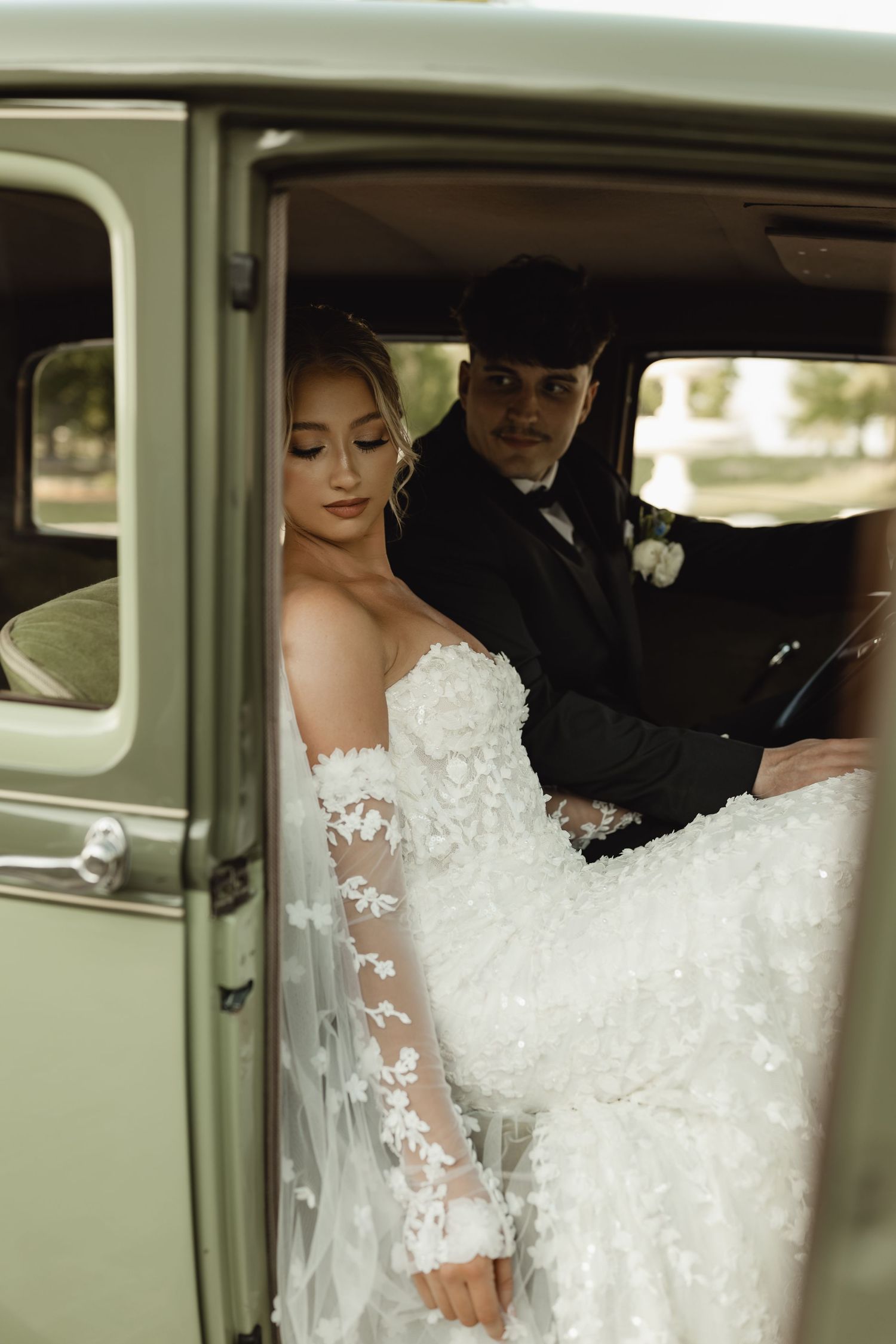 Bride in lace gown stepping out of vintage green car with groom in black suit during wedding day.