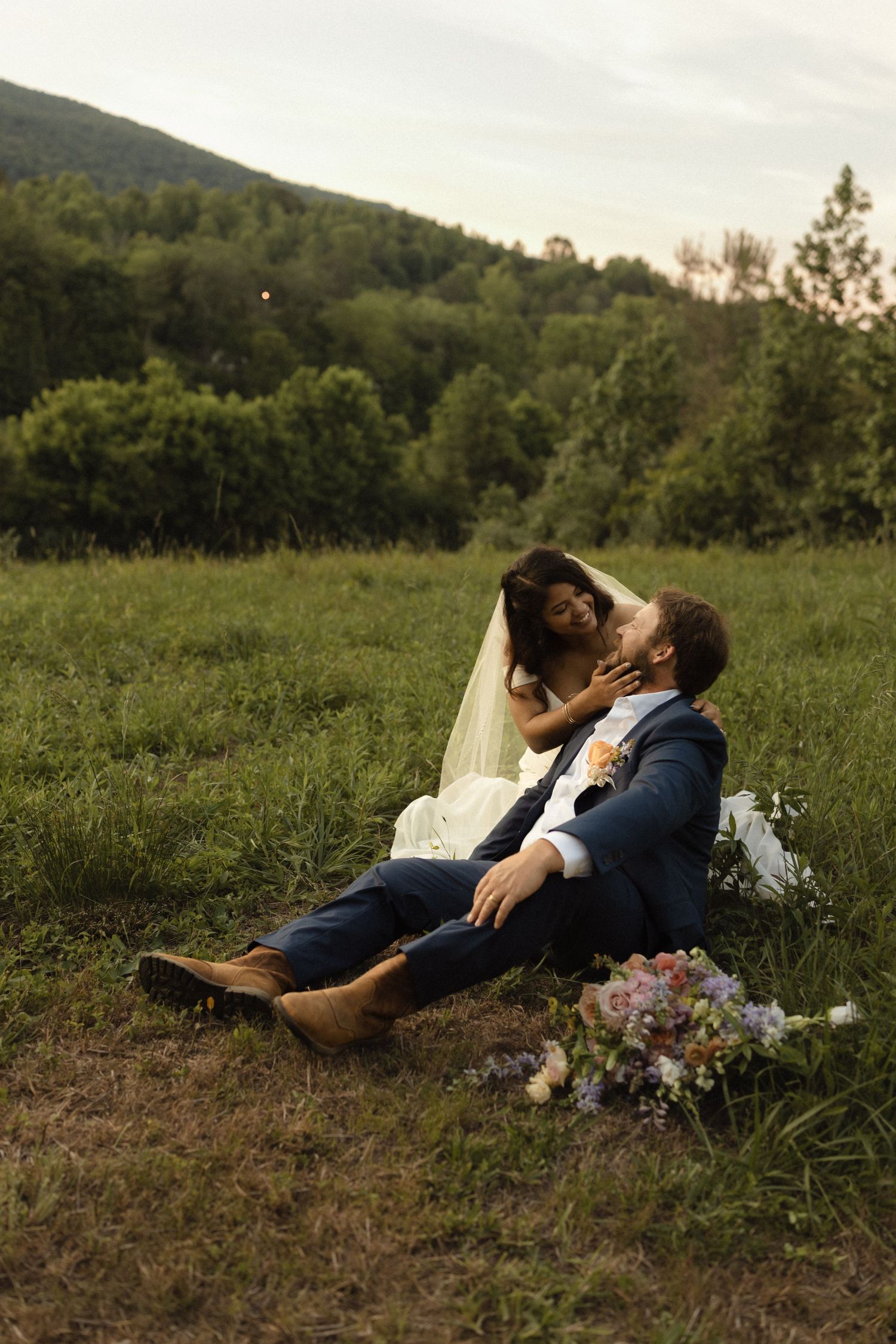 Bride and groom sitting together in a lush green field at dusk, with mountains and wildflowers nearby.