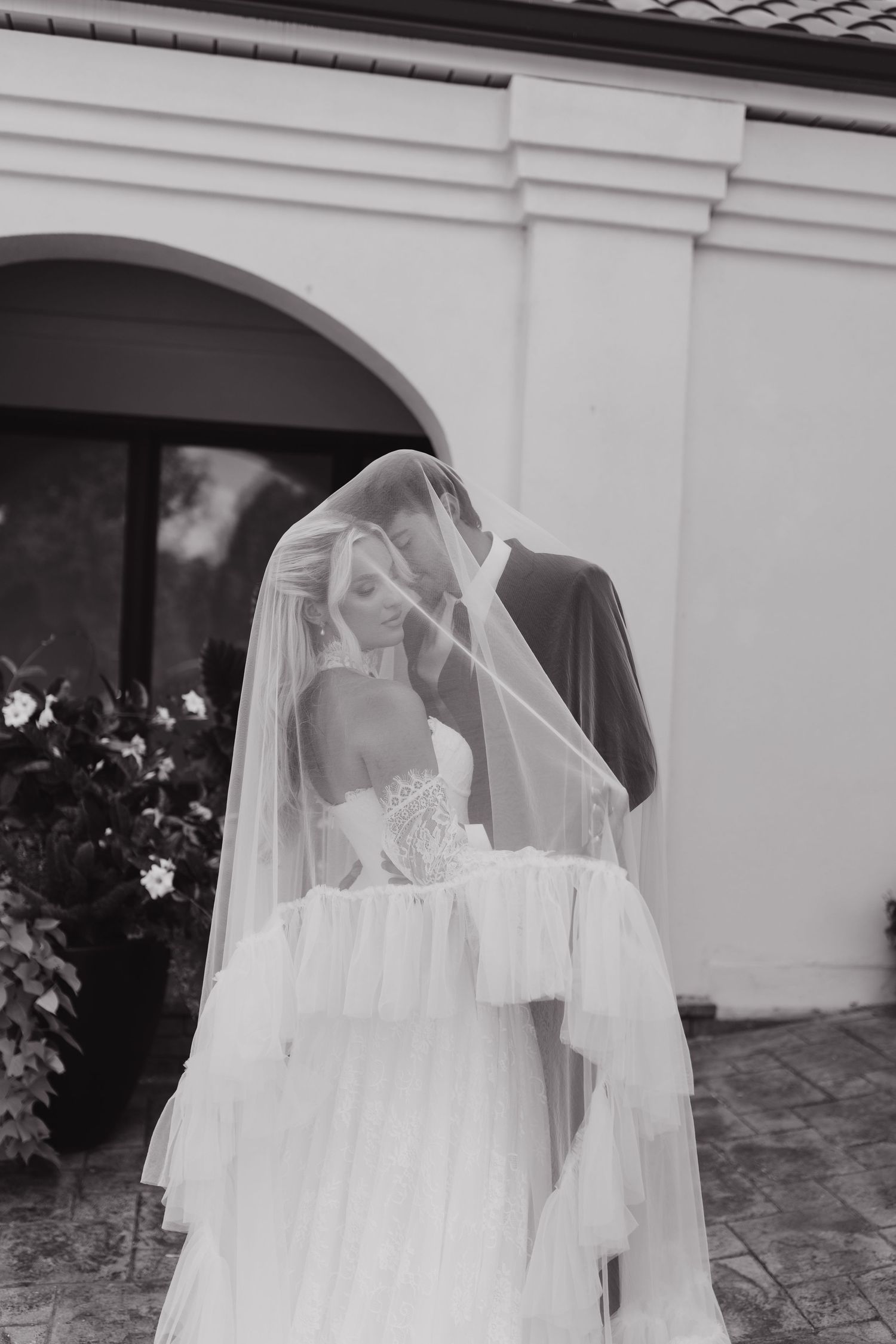 Black and white photo of bride and groom sharing a kiss under a flowing veil outside an elegant venue.