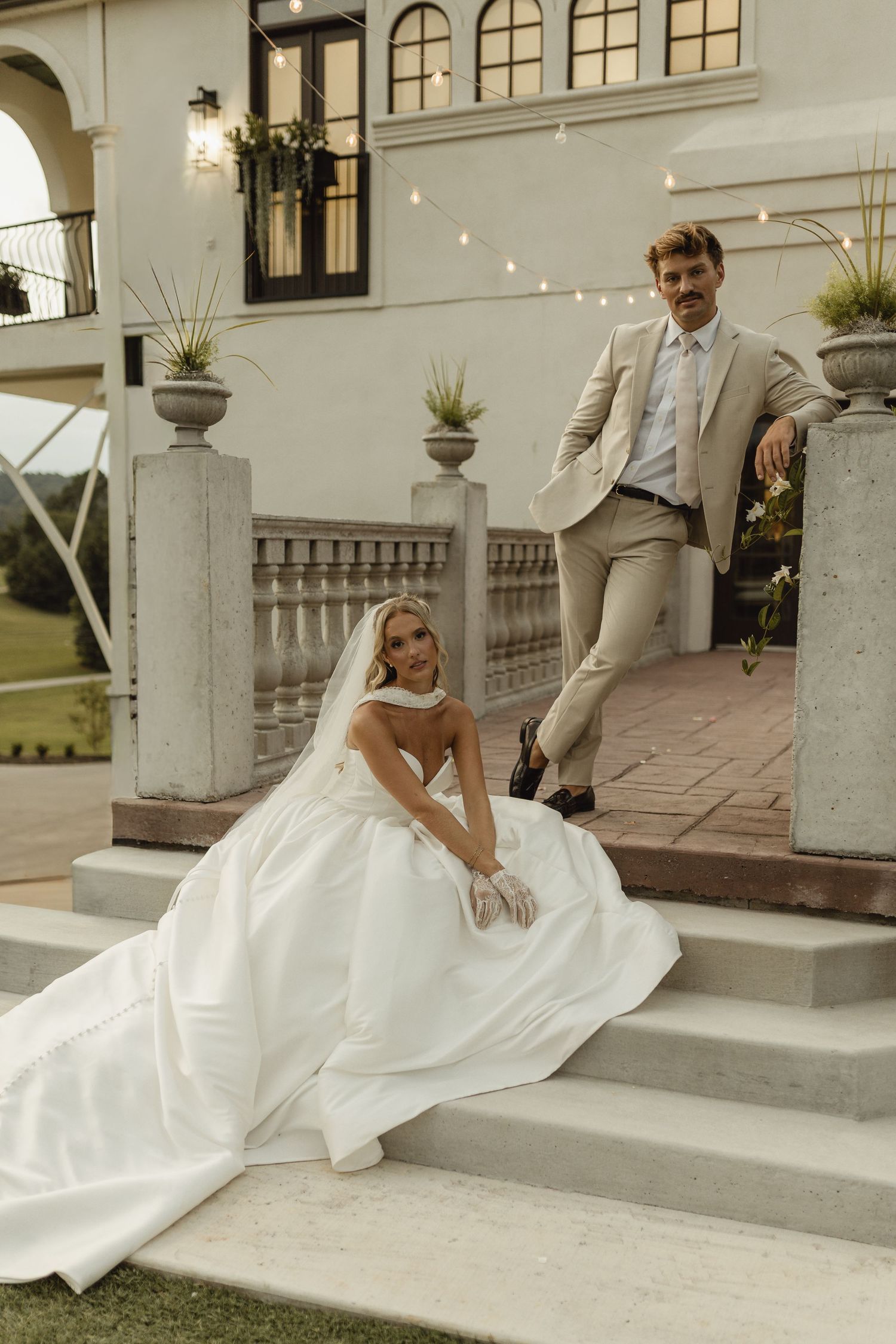 Bride in white ball gown sits on stone steps beside groom in tan suit at elegant venue with string lights.