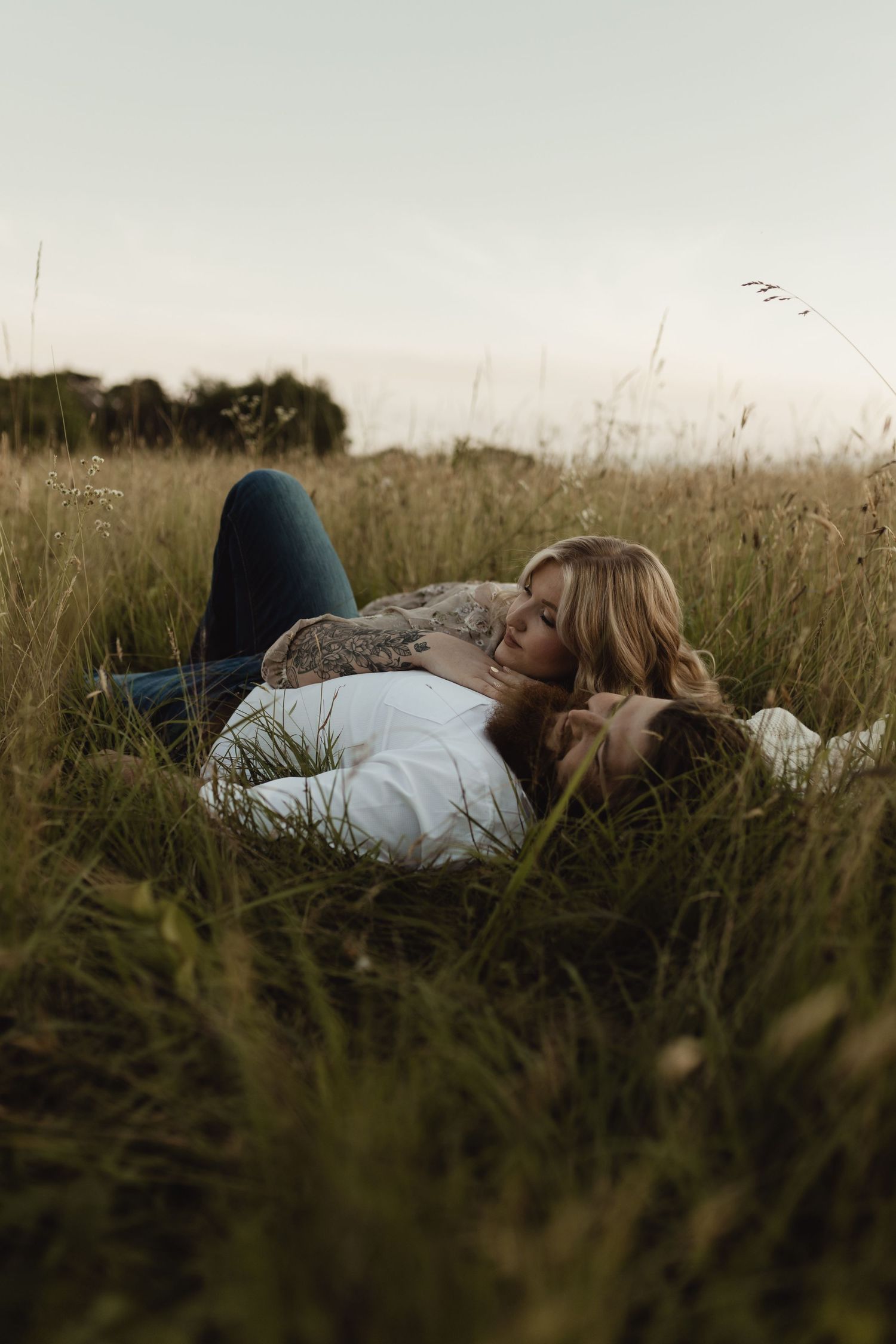 Couple lying together in tall grass field, sharing a romantic moment outdoors in a serene meadow setting.
