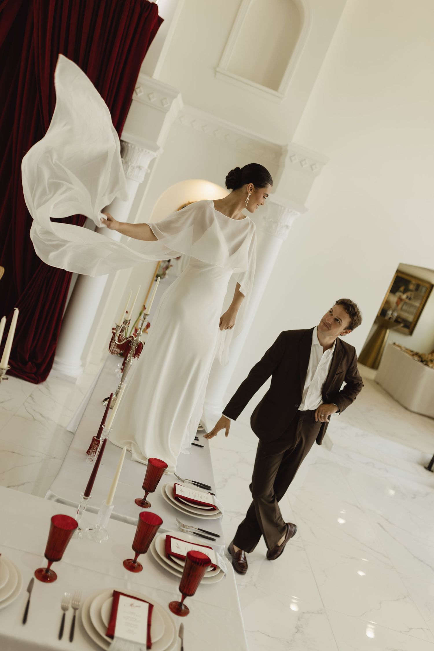 Bride in white dress and groom in dark suit stand near elegantly set table with red goblets in white hall.