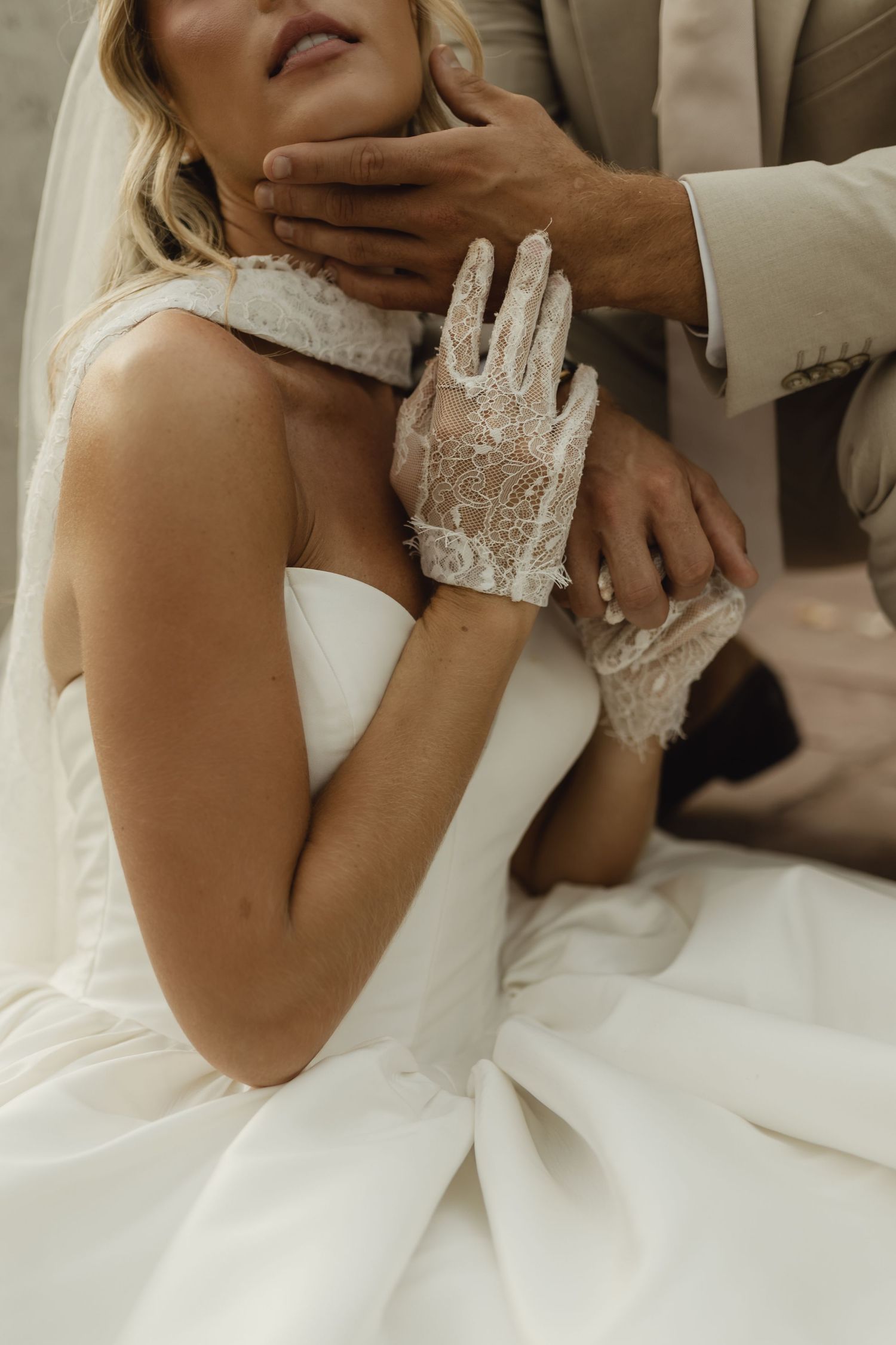 Bride in white strapless gown and lace gloves with groom in tan suit tenderly holding her face at wedding.