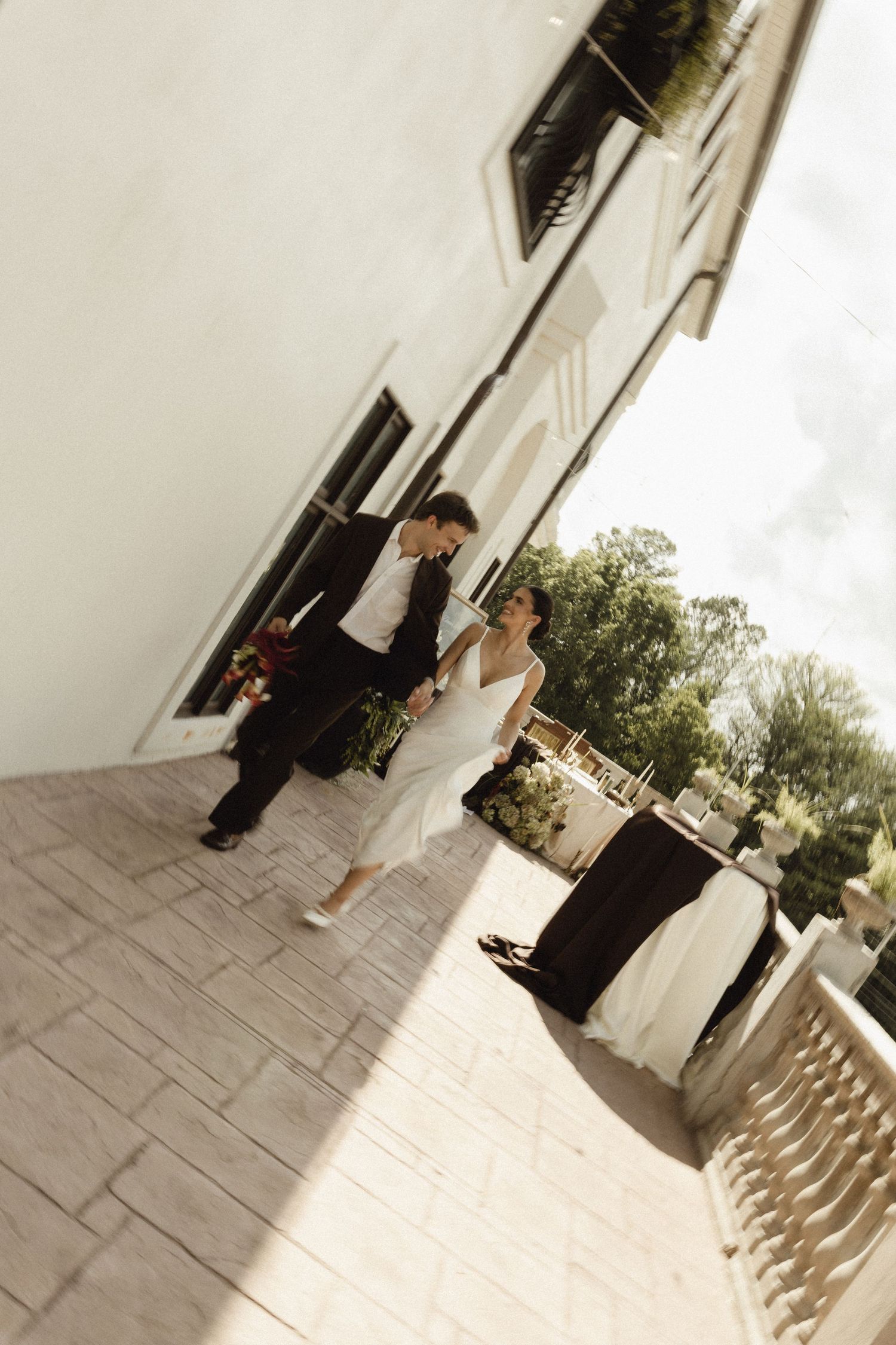 A bride and groom walk hand in hand on a sunlit terrace outside an elegant white building.