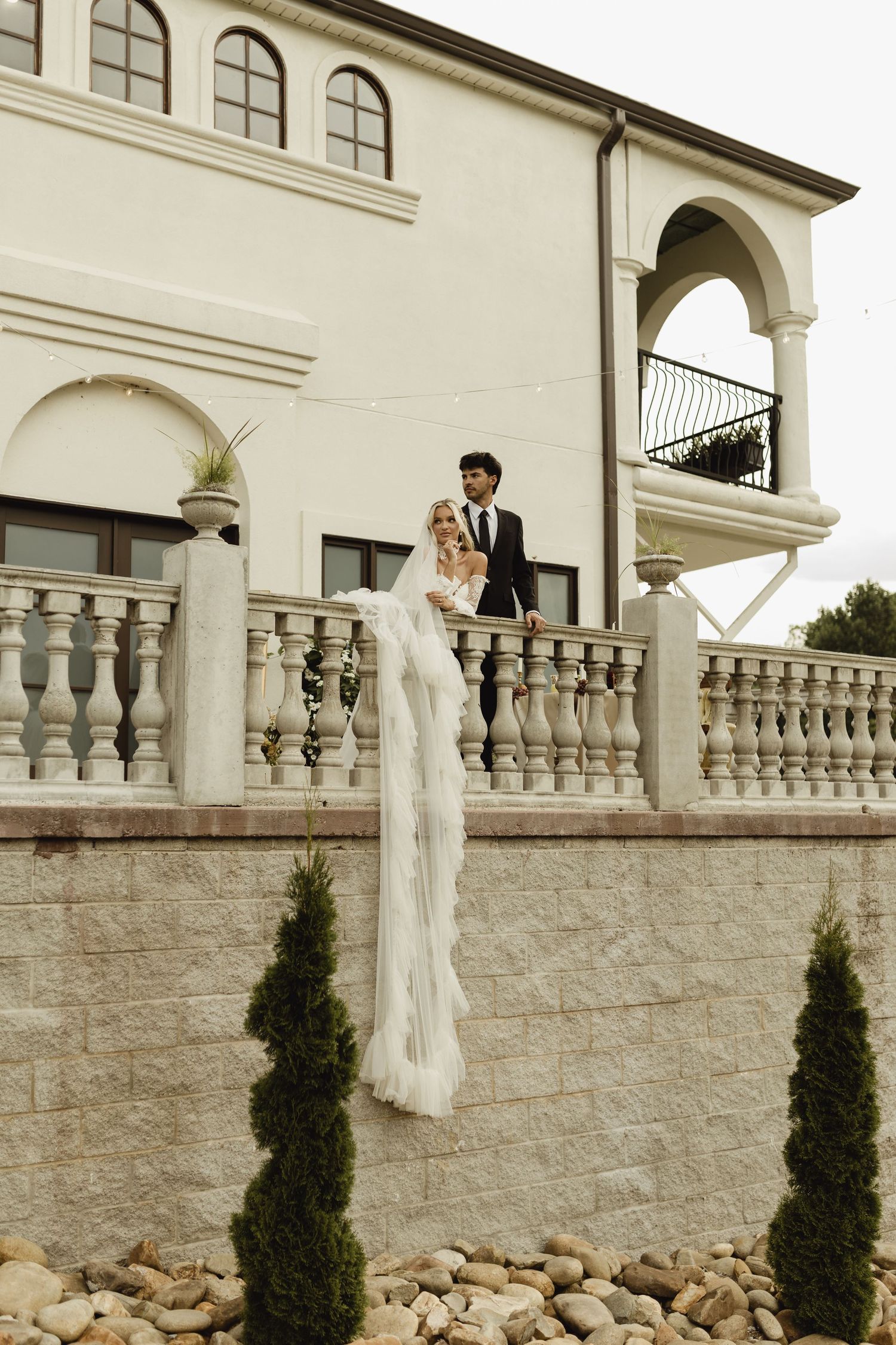 Bride in white gown with long veil and groom in black suit posing on elegant balustrade of white villa.