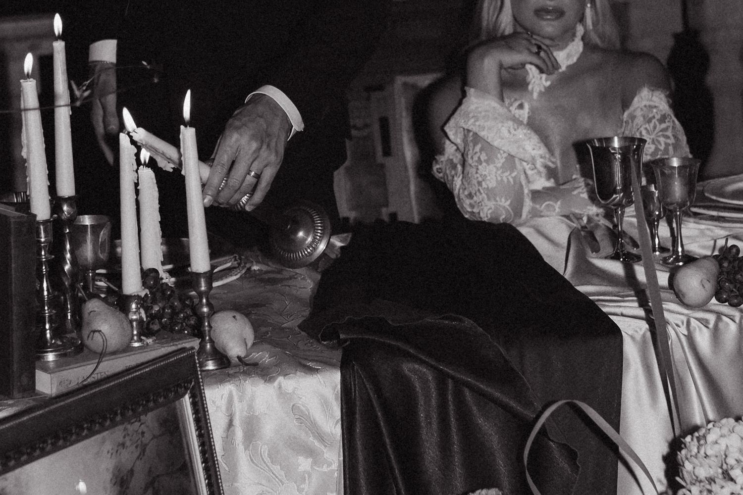 Black and white photo of elegantly dressed people at a candlelit table with goblets and fruit.