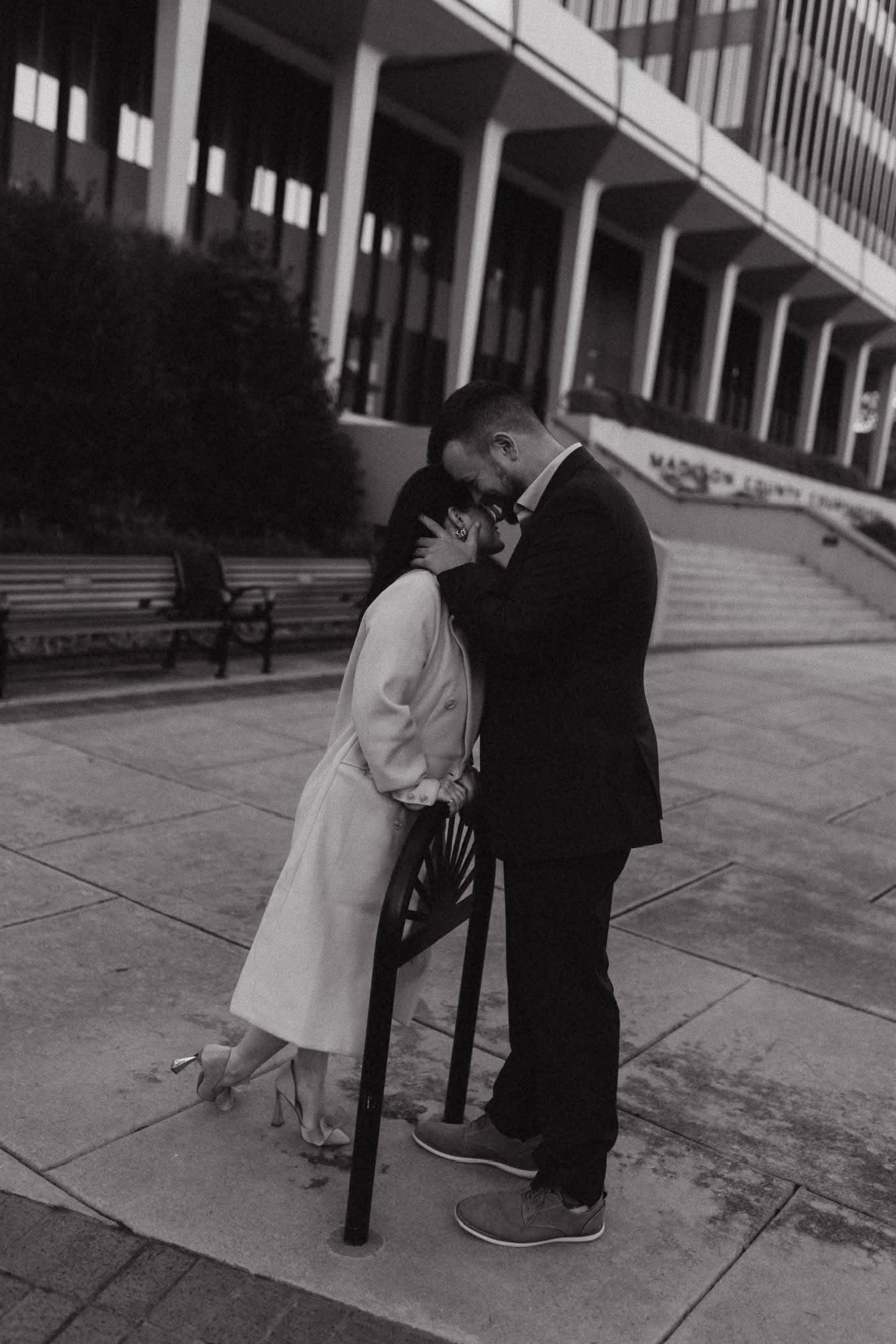 Black and white photo of a couple sharing an intimate moment outside a building, the woman in a white dress leaning into the man.