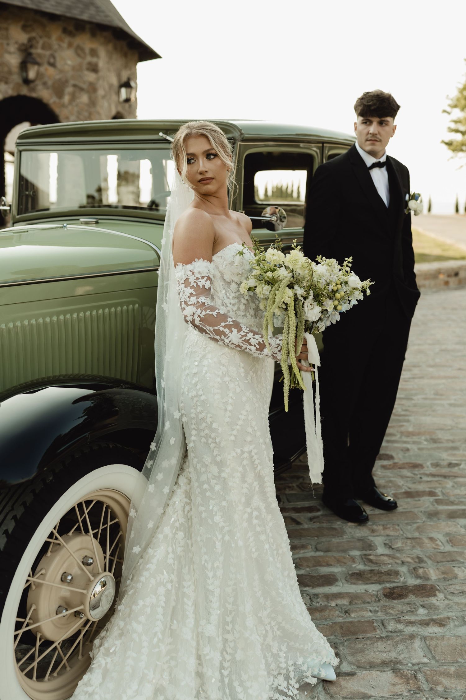 Bride in lace mermaid gown with veil holds bouquet beside vintage green car as groom in black tuxedo stands behind.