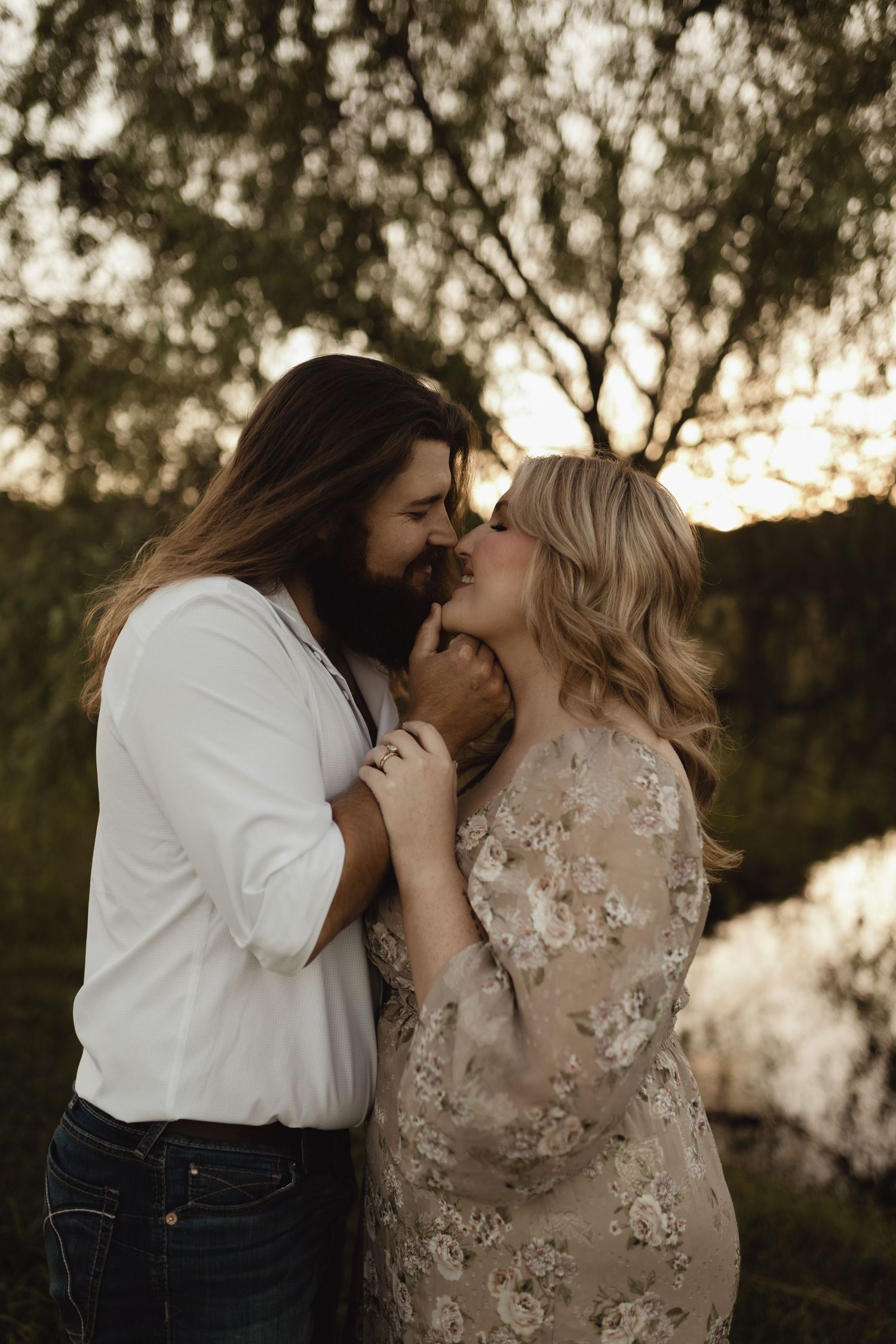 A bearded man and blonde woman in floral dress share a romantic moment at golden hour outdoors.