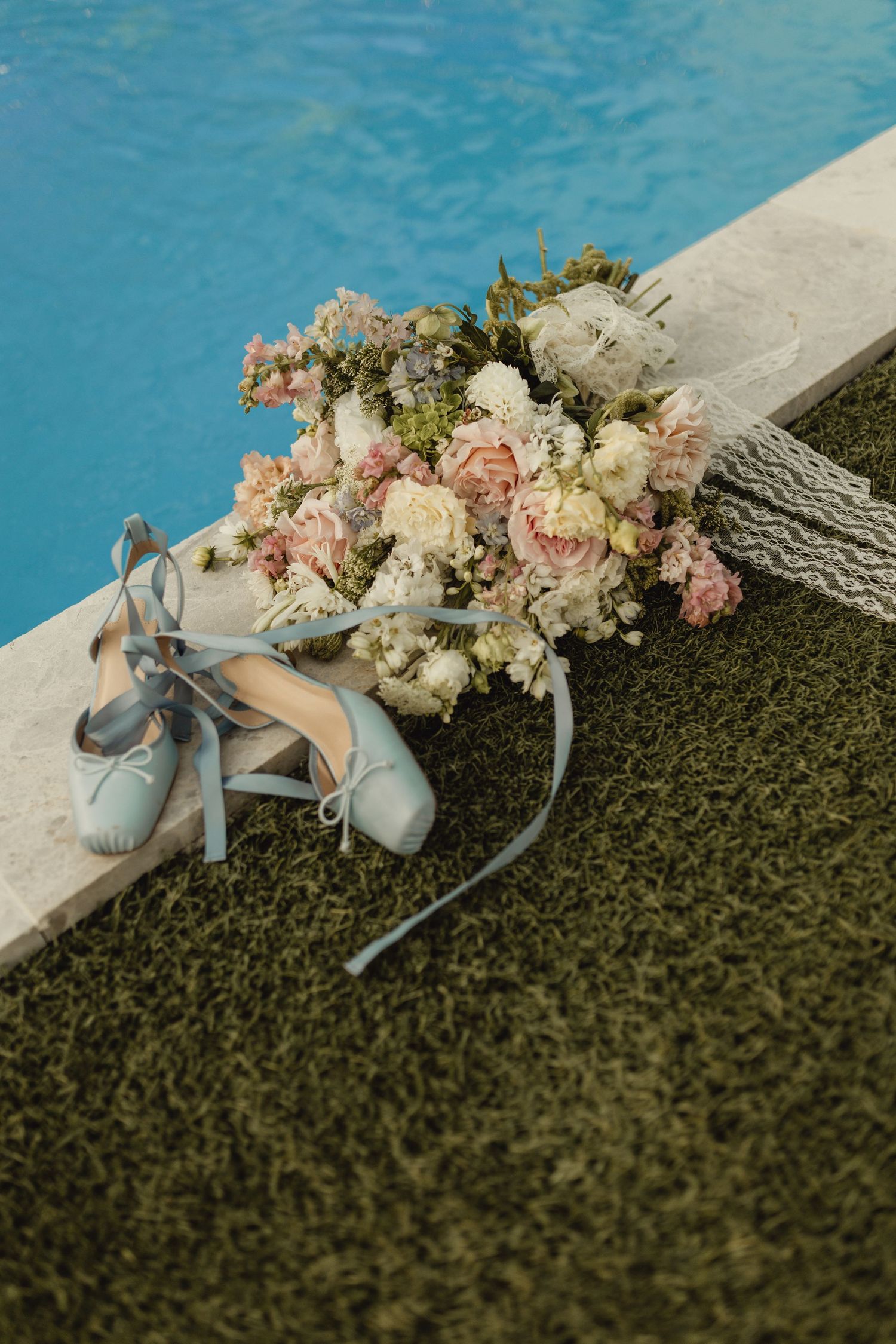 Light blue bridal ballet flats and pink floral bouquet with ribbon resting on grass beside a swimming pool.