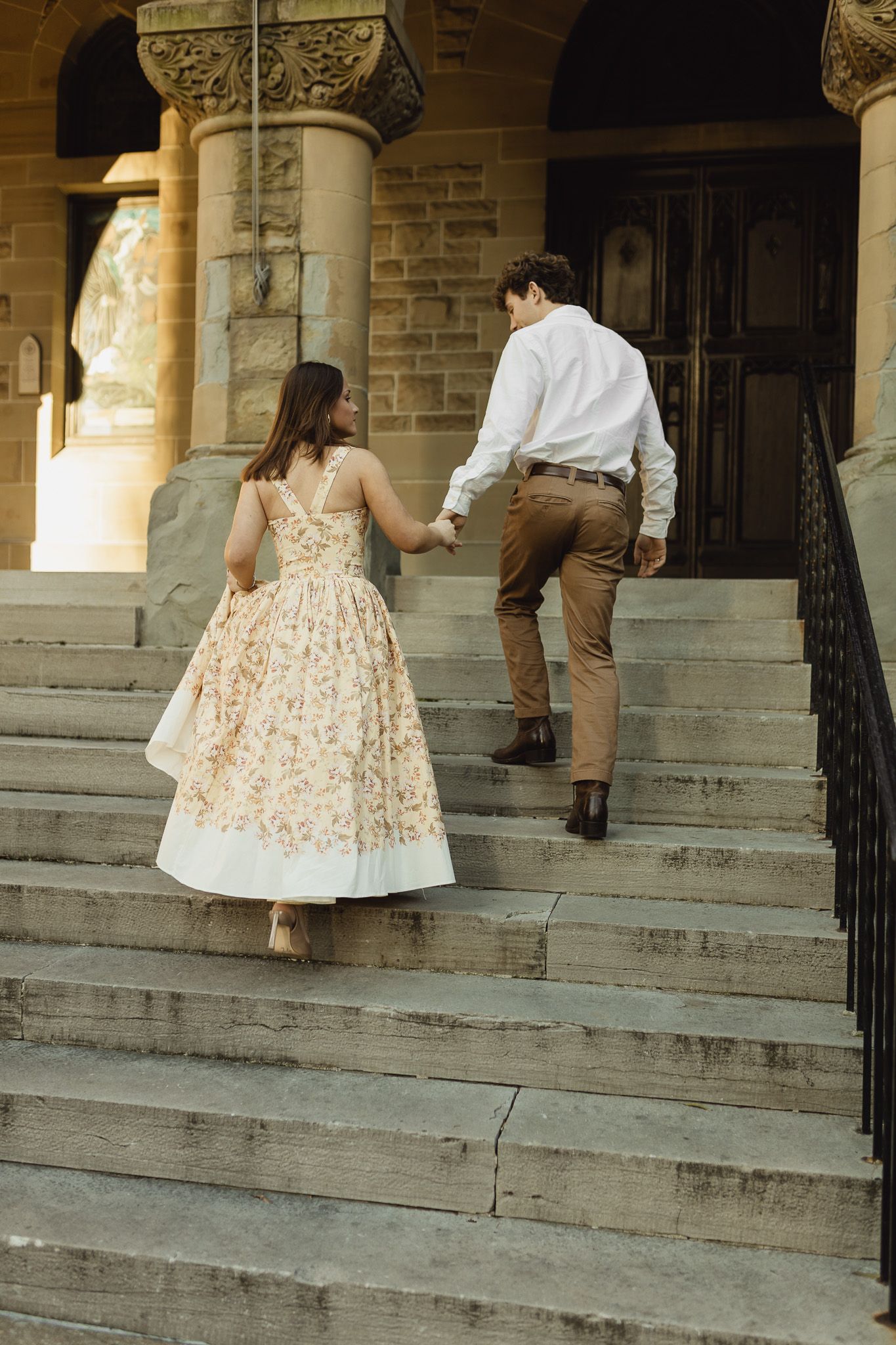 Couple holding hands walking up stone steps of a historic building, woman in floral midi dress.