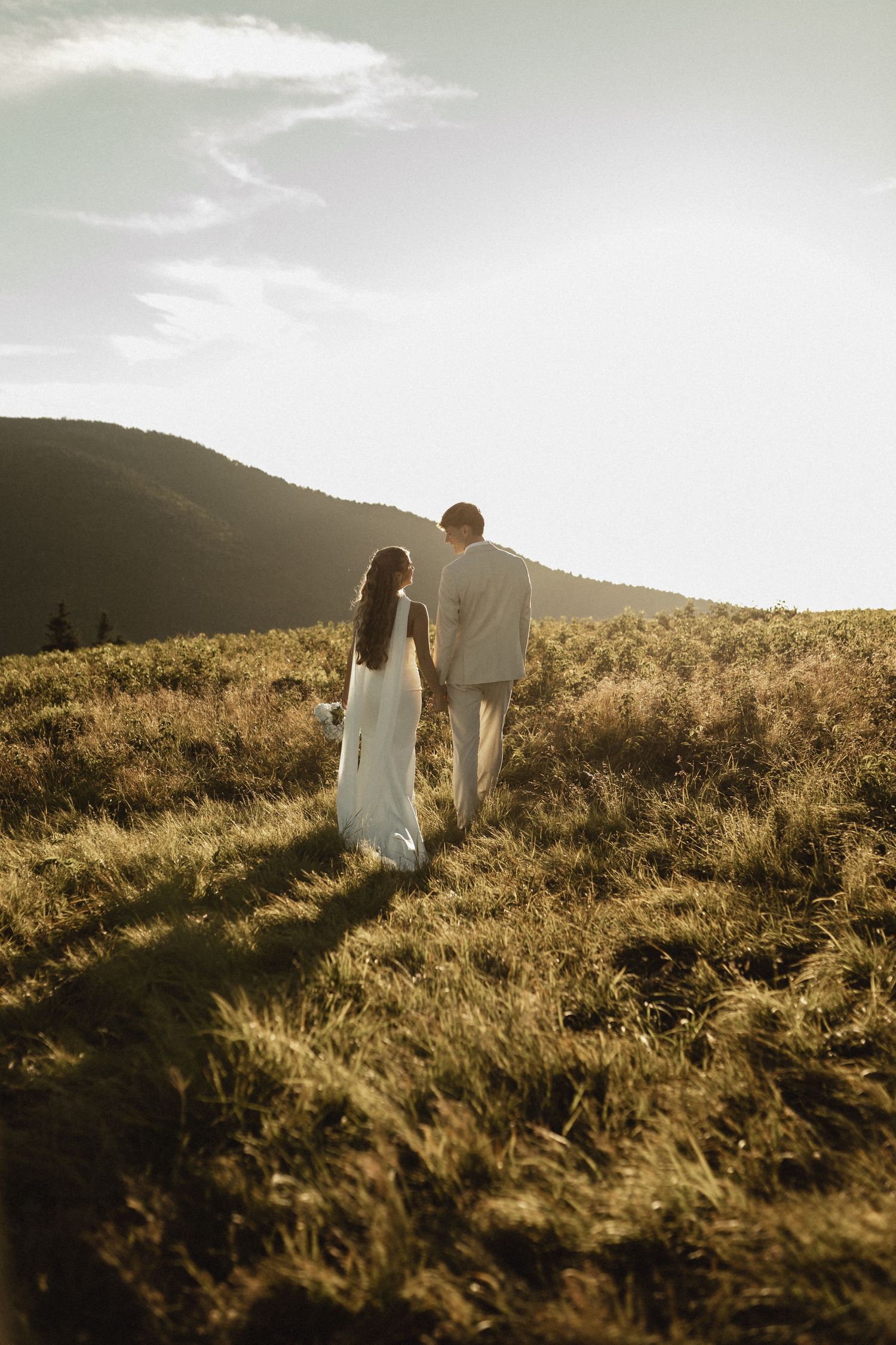 Couple in wedding attire holding hands walking through golden meadow with mountain backdrop at sunset.