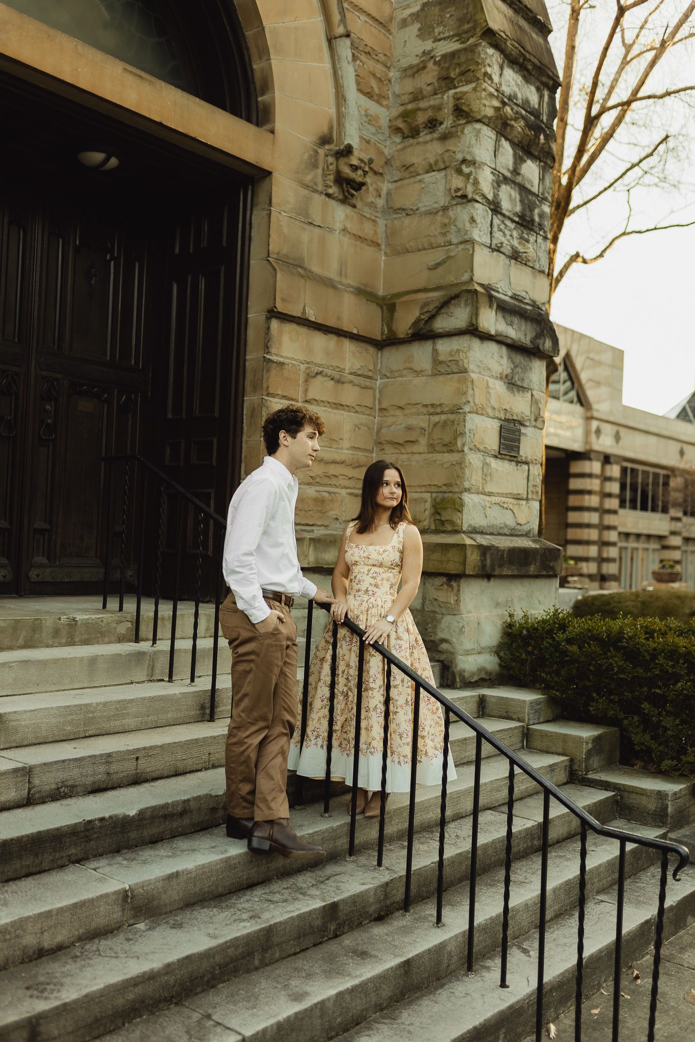 A couple stands on stone steps outside a historic building, the woman in a floral dress and man in white shirt and tan pants.