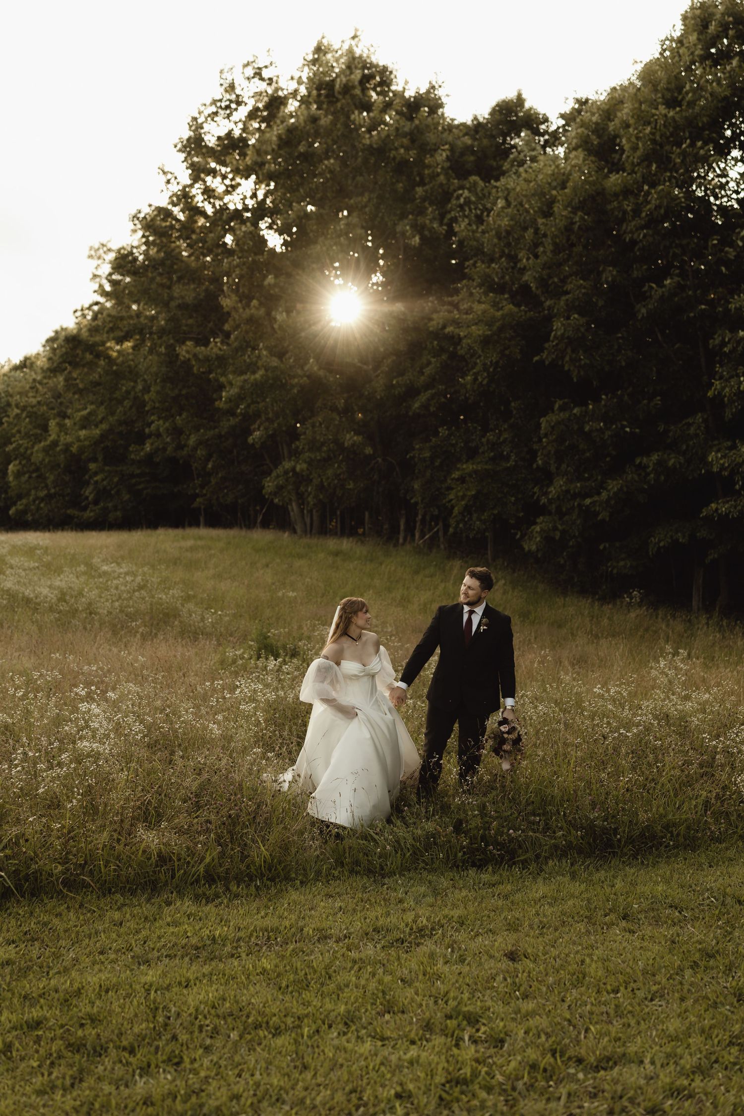 Bride and groom walking hand in hand through a sunlit meadow with lush green trees at golden hour.