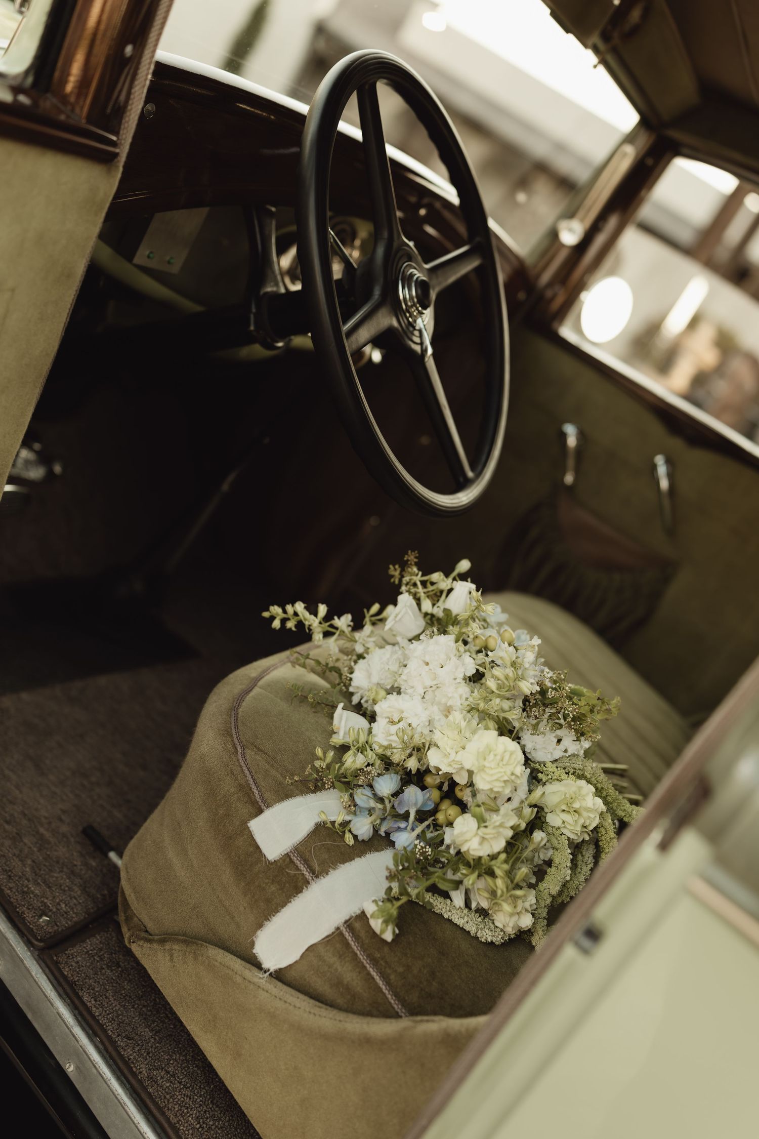 A bridal bouquet of white flowers rests on the seat of a vintage classic car near the steering wheel.