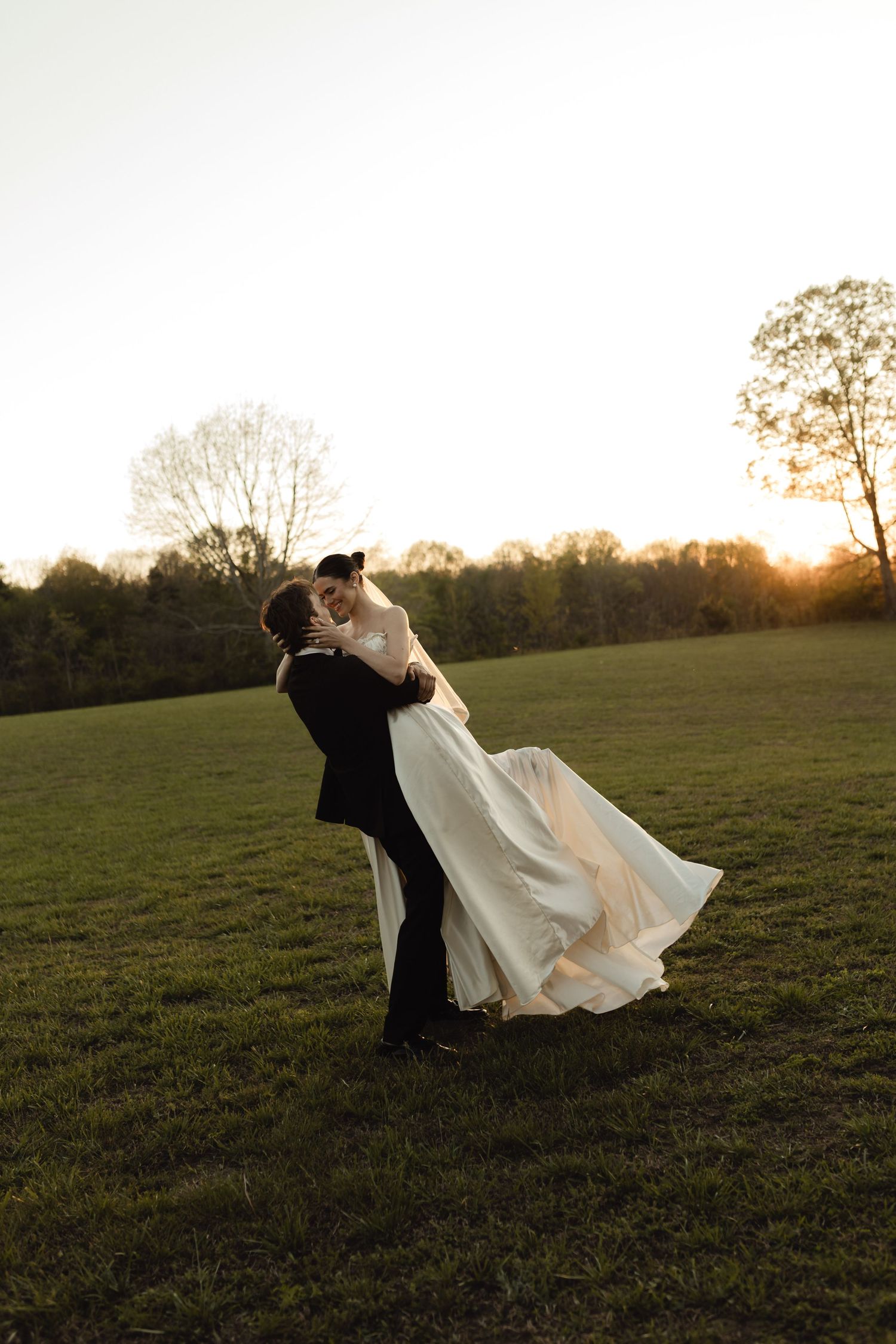 Groom lifting bride in white flowing gown during golden hour sunset on lush green field.