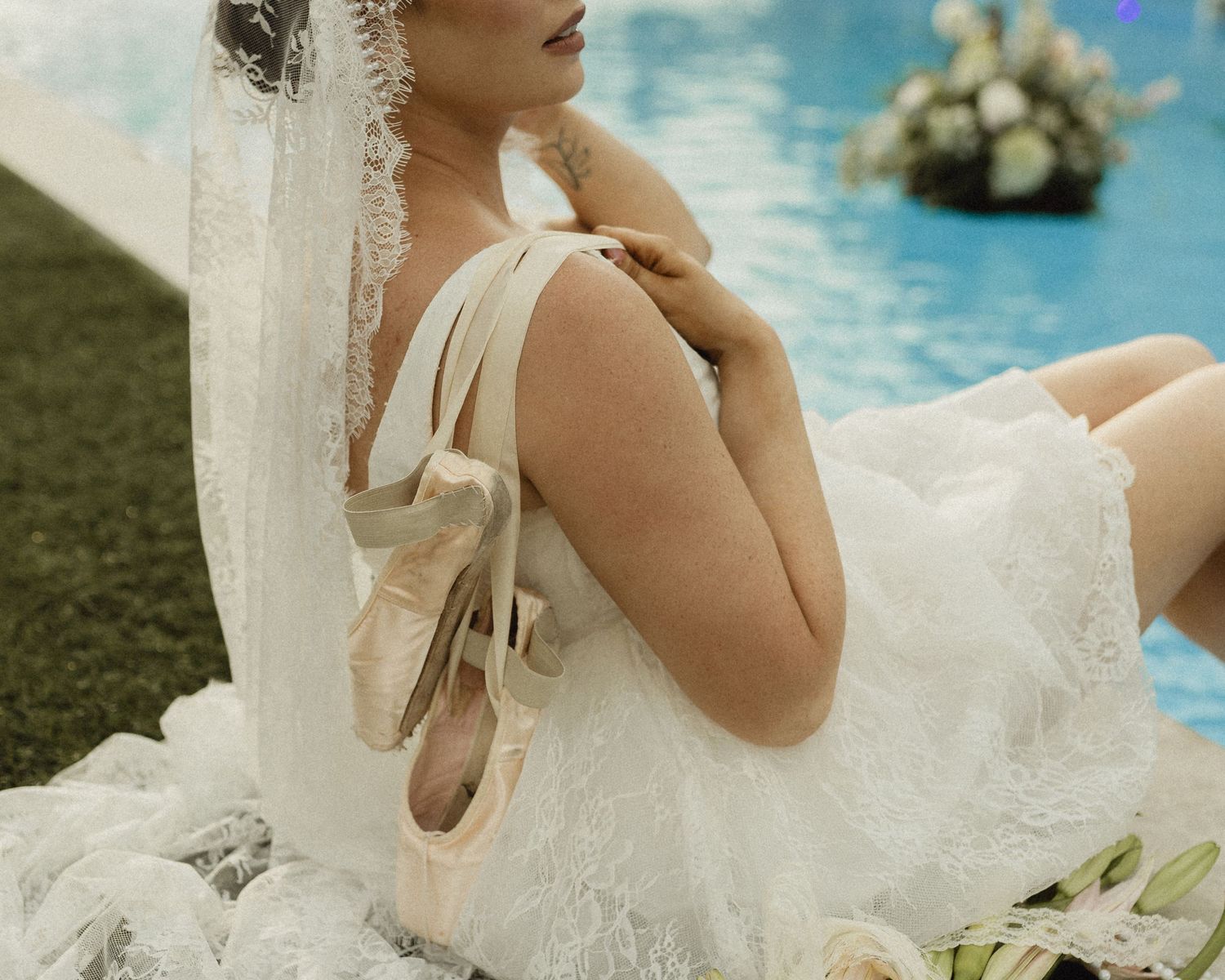 Ballerina in white lace dress holding pointe shoes beside a pool with floral arrangements in background.