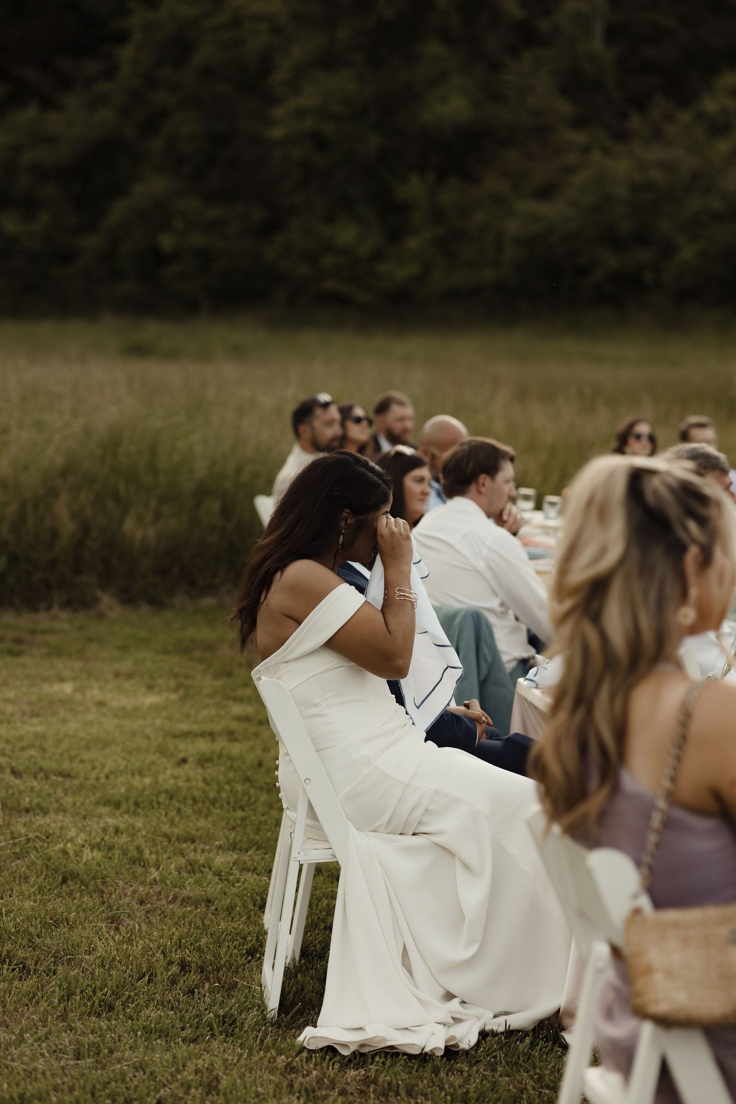 Outdoor wedding reception with guests seated at long tables on a grass field, bride in white off-shoulder gown in foreground.