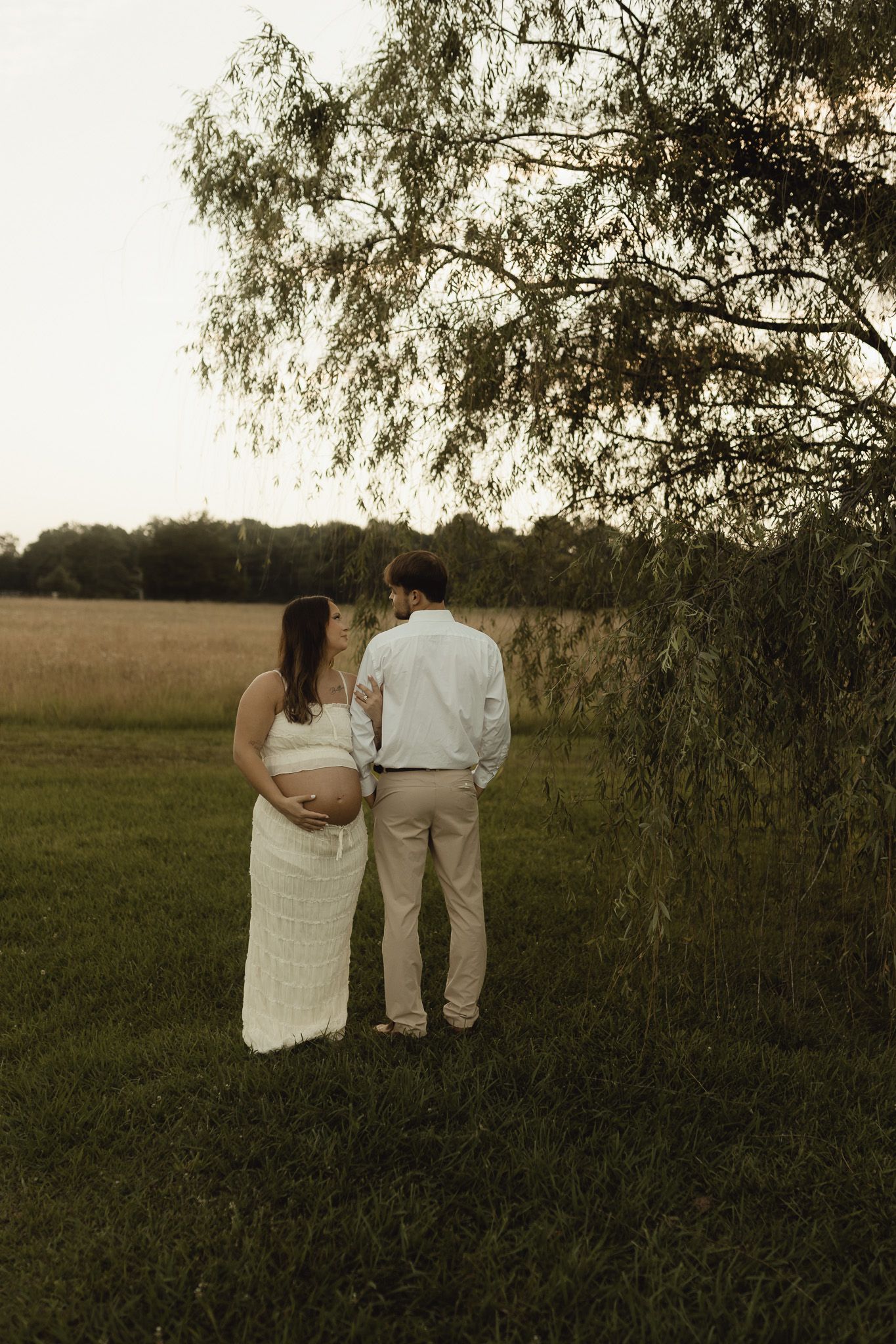 Pregnant woman in white lace dress and man in white shirt stand under willow tree overlooking green field at dusk.