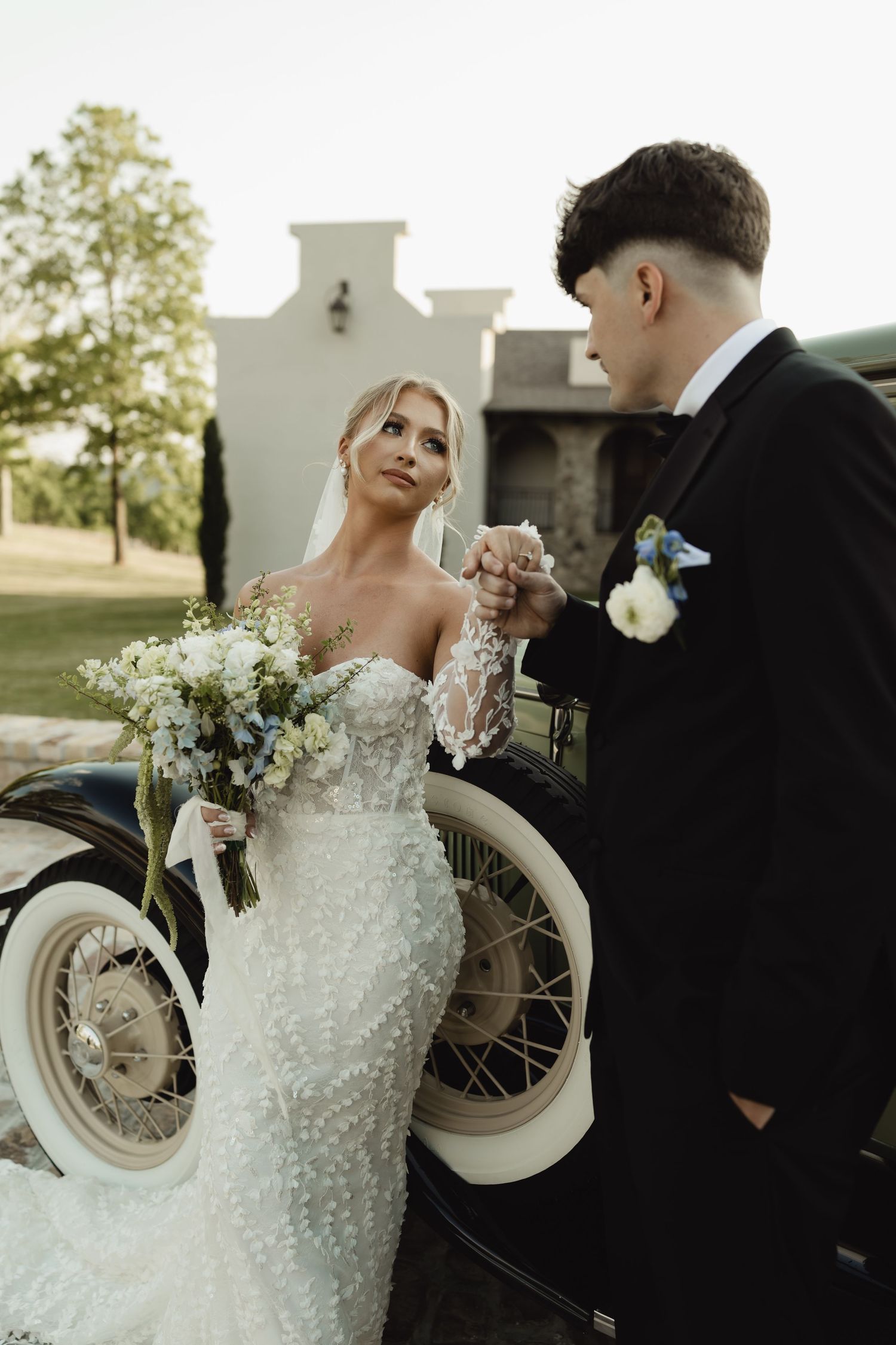 Bride in lace mermaid gown holds bouquet as groom in black tuxedo holds her hand beside vintage car outdoors.