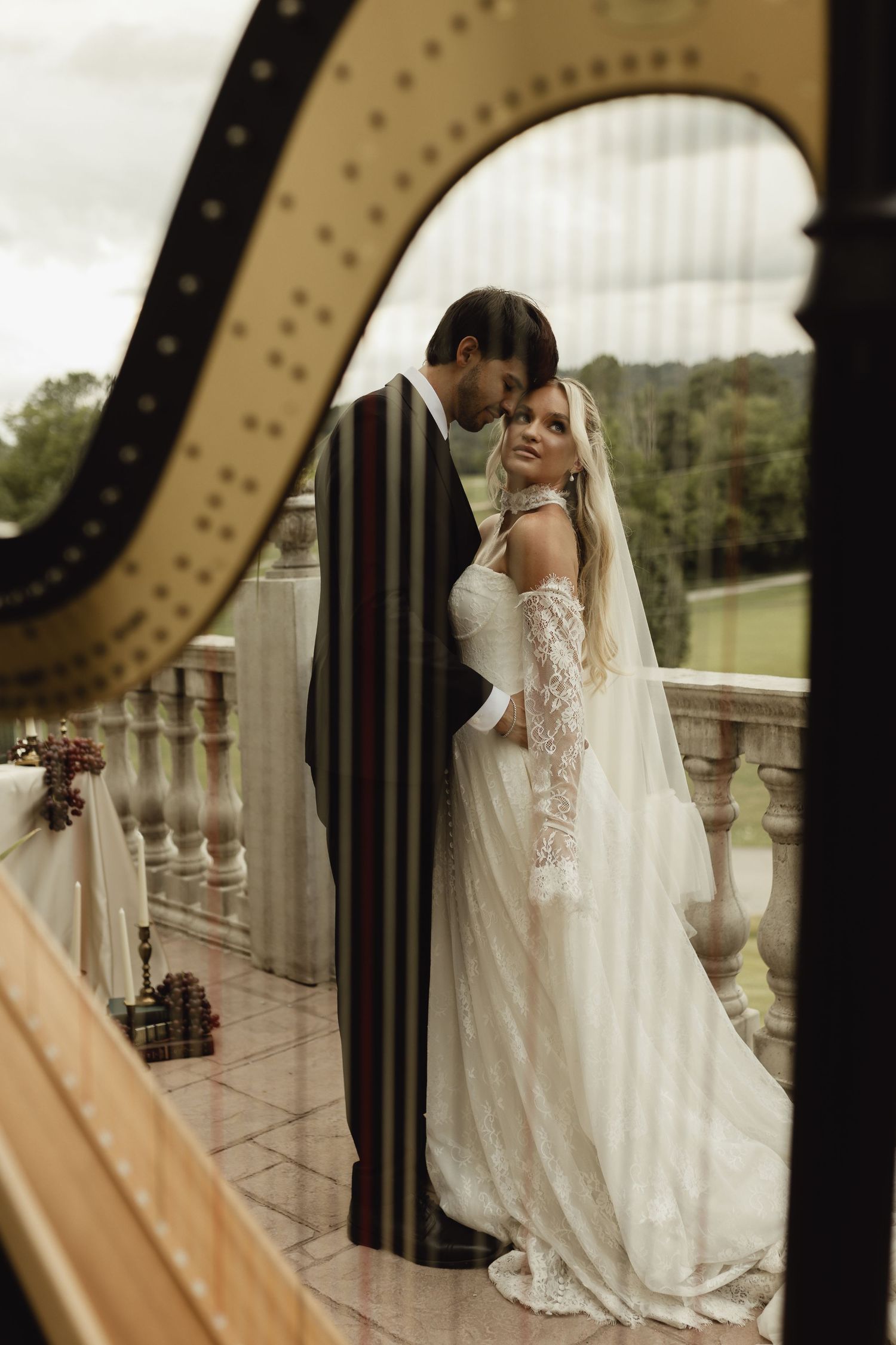 A bride and groom share a romantic moment on a balcony, framed through the strings of a golden harp.