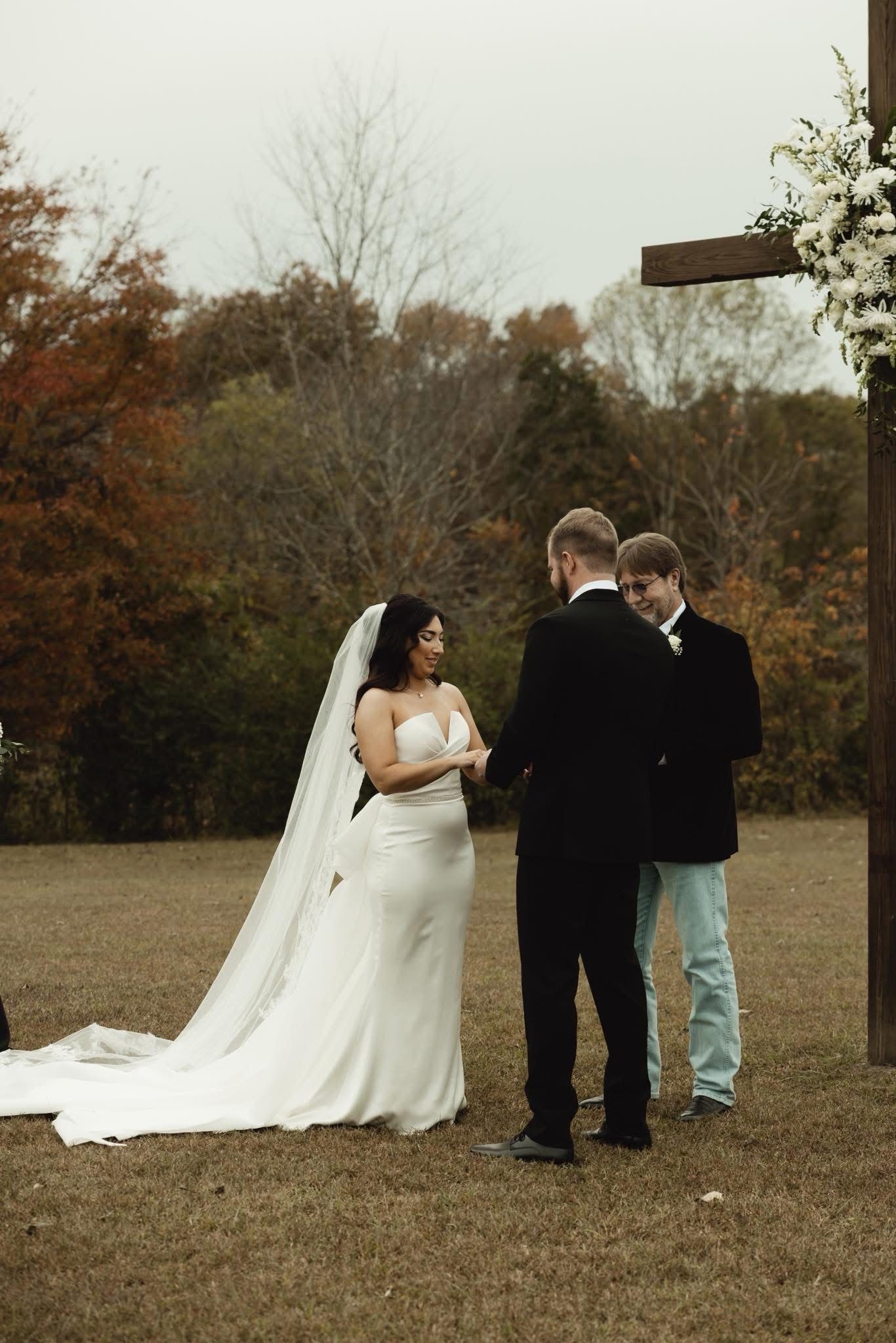 Outdoor fall wedding ceremony with bride in white gown and veil, groom in black suit, exchanging vows with officiant.