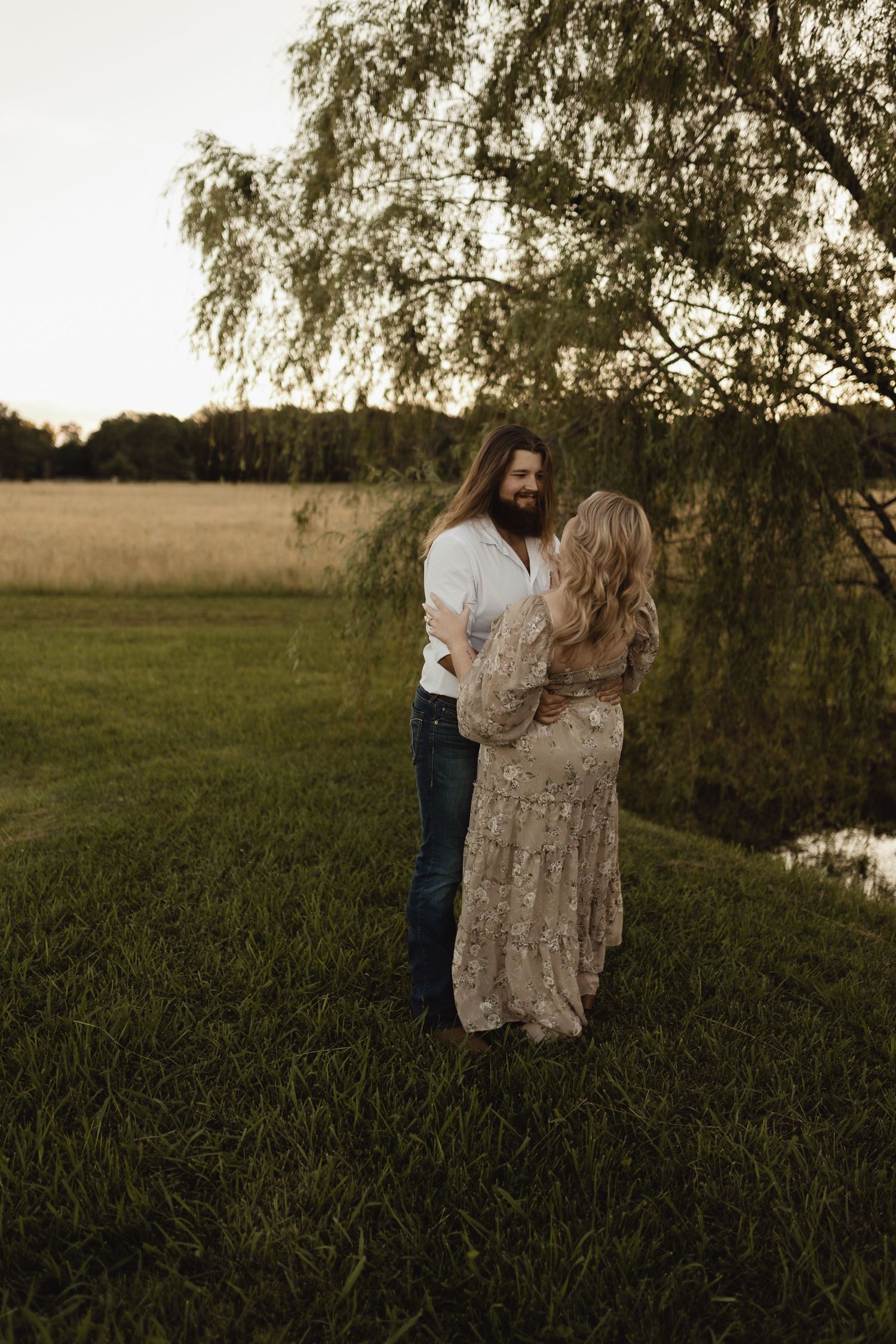 Couple embracing at golden hour near a pond, woman in lace dress, man in white shirt and jeans, under willow tree.