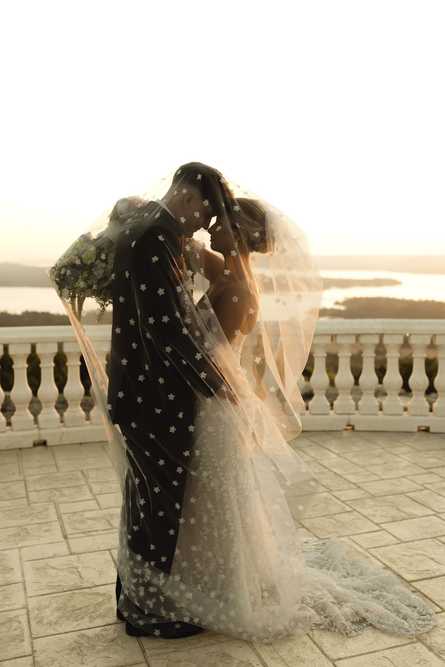 Bride and groom share romantic kiss under polka dot veil on scenic waterfront terrace at golden hour sunset.