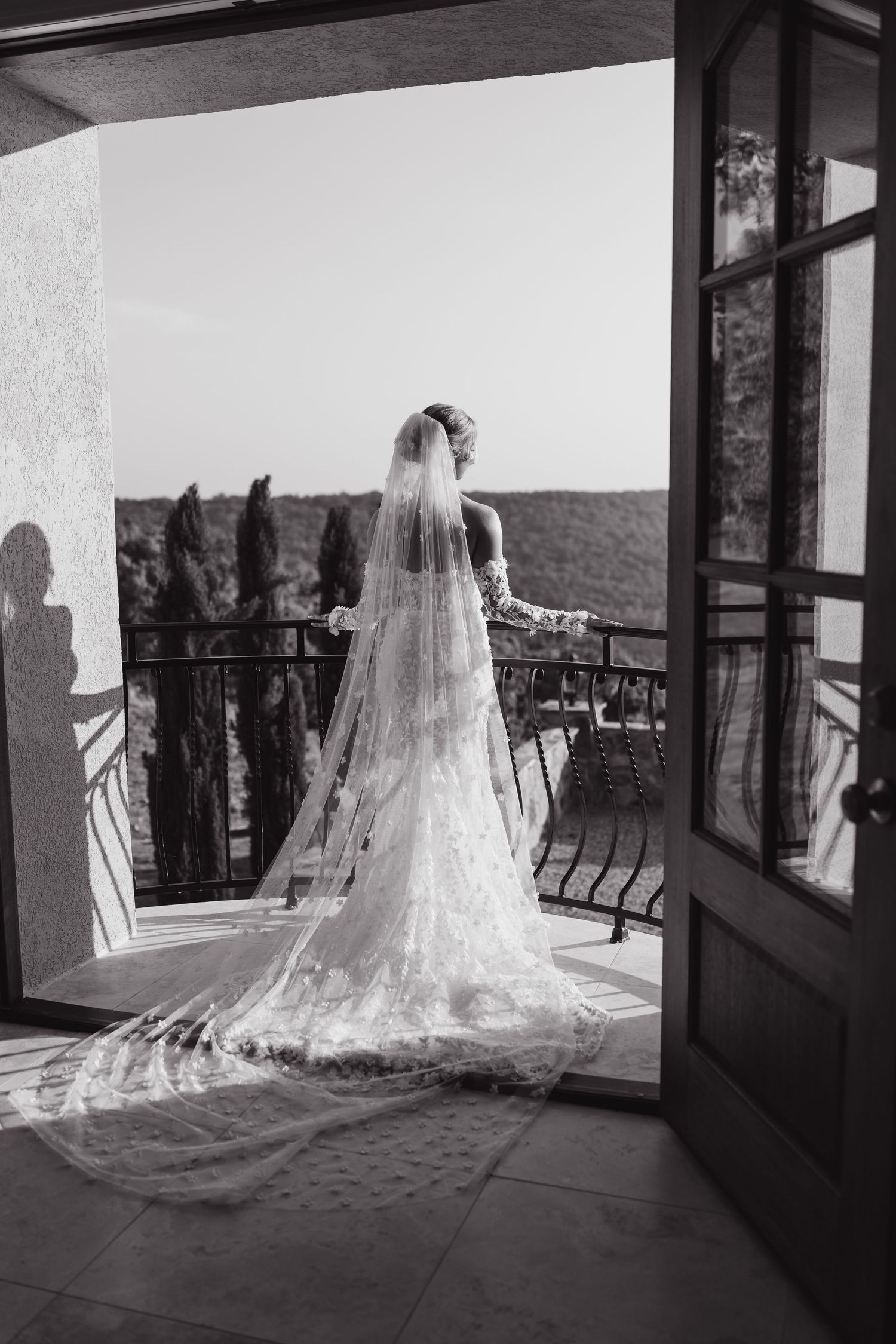 Bride in lace wedding gown and long veil stands on balcony overlooking scenic landscape, black and white photo.