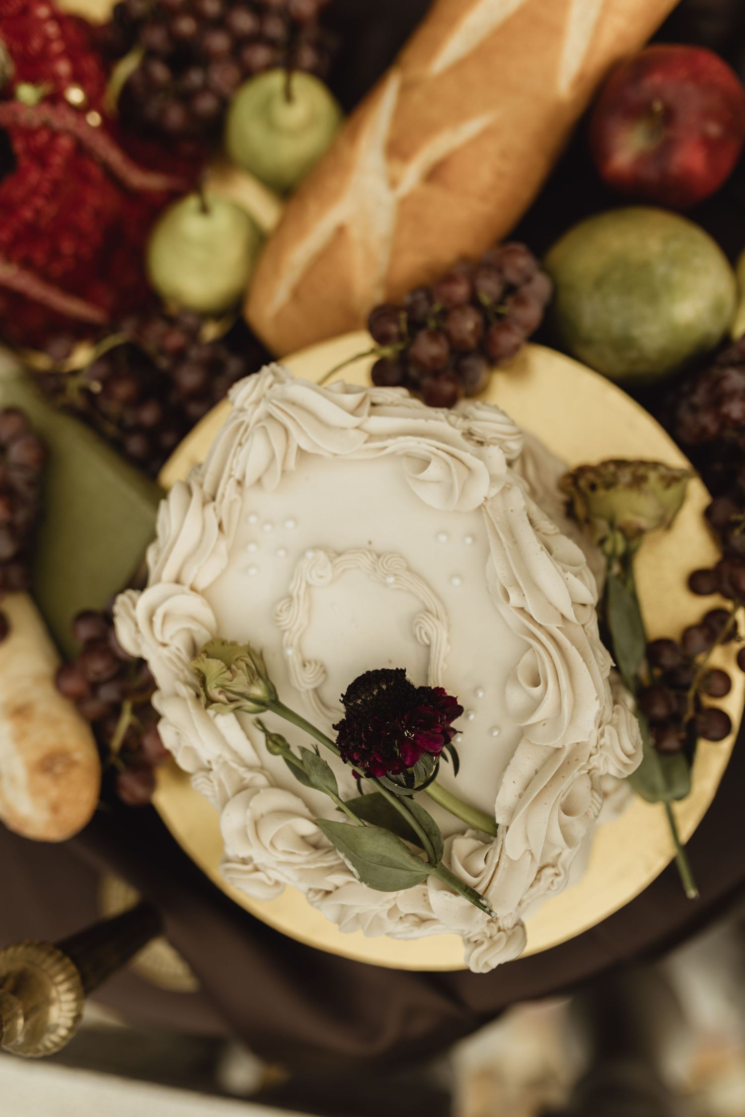 Elegant white frosted cake topped with a dark red rose, surrounded by grapes, pears, pomegranate, and a baguette.