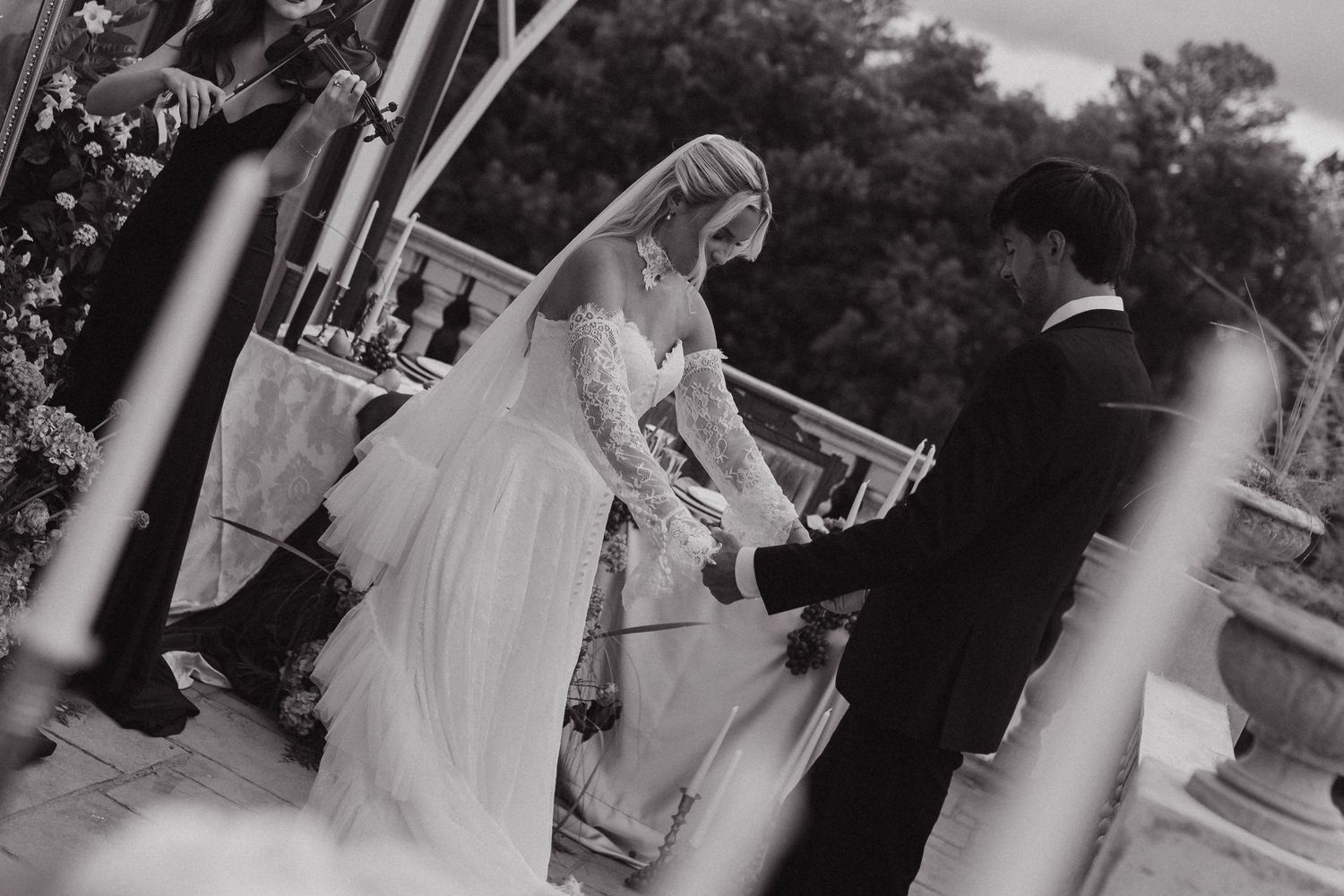 Black and white wedding photo of bride in lace gown and groom holding hands during outdoor ceremony.