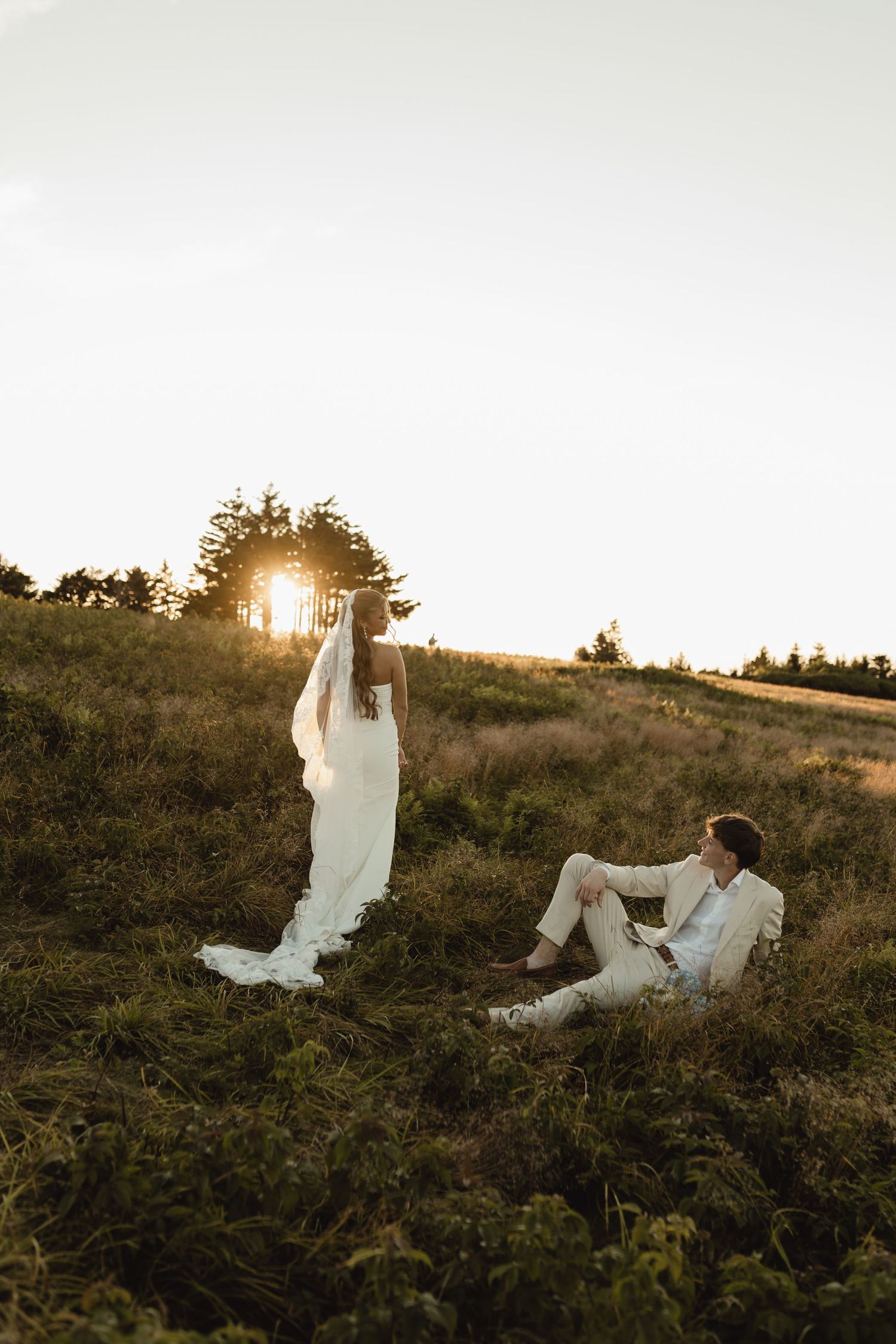 Bride and groom in white attire on a sunlit grassy hill at golden hour sunset with trees in background.