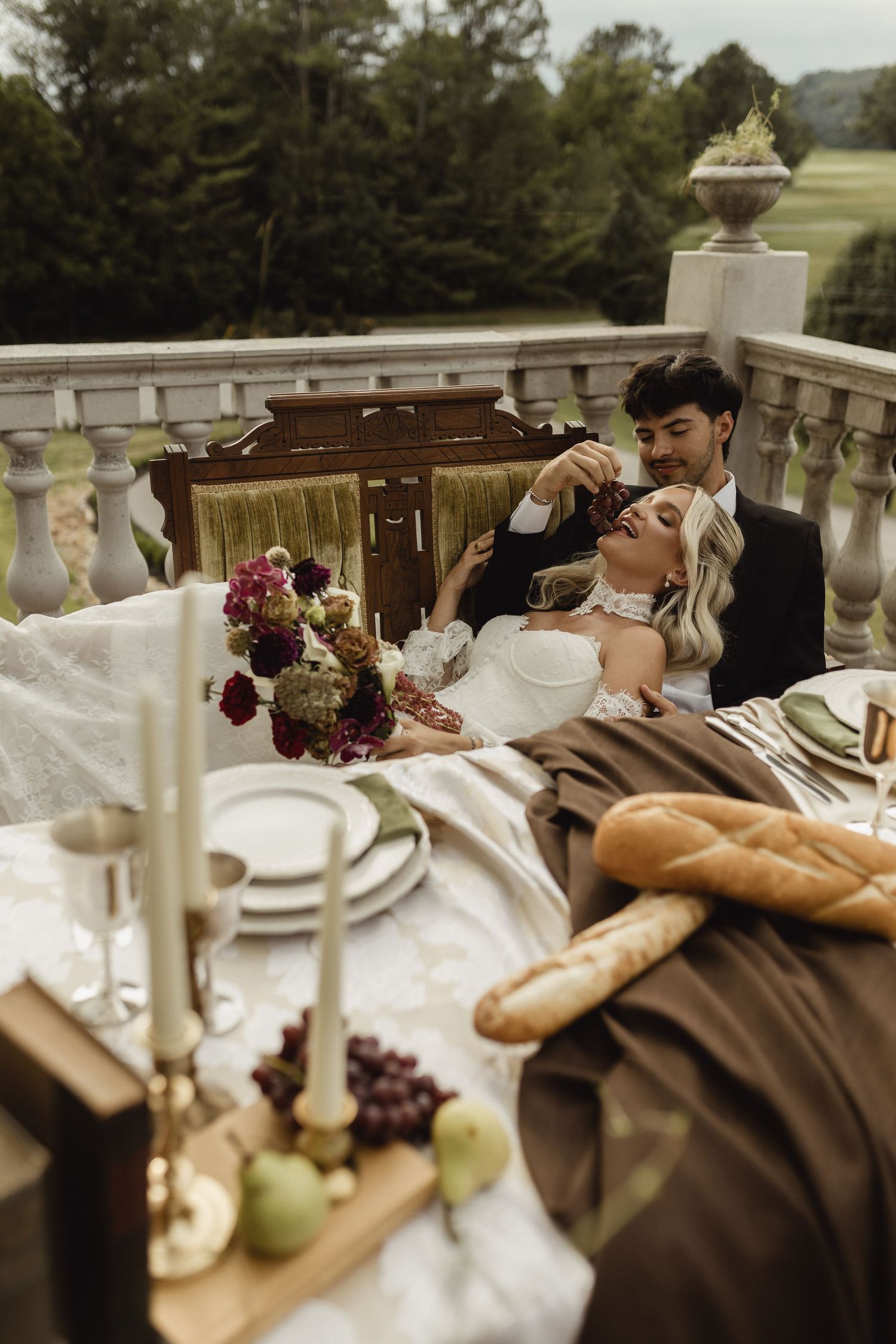 A bride and groom share an intimate moment at an elegant outdoor tablescape with florals, candles, bread, and grapes on a stone balcony.