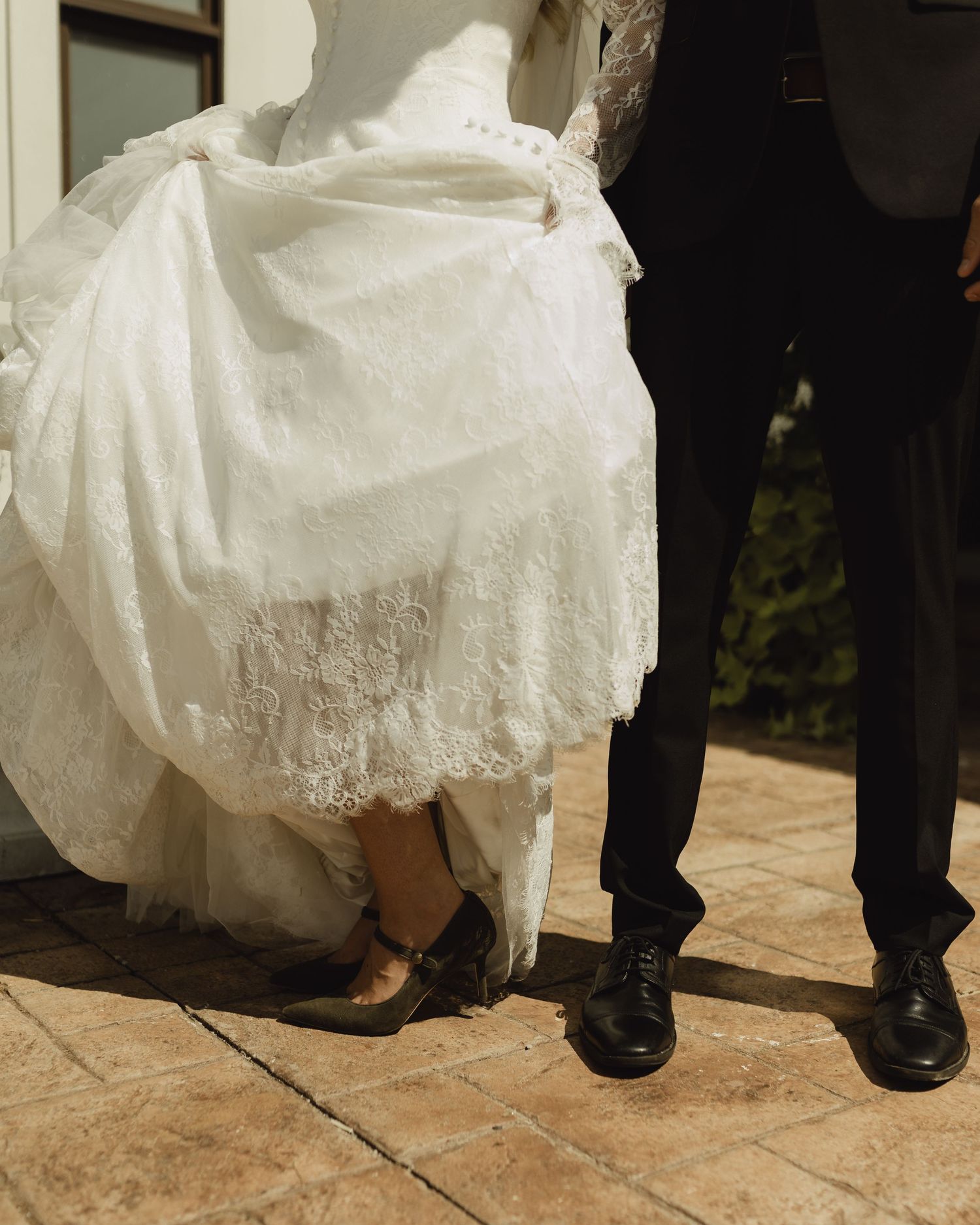 Close-up of bride in lace wedding gown and groom in dark suit standing on stone patio, showing elegant footwear details.