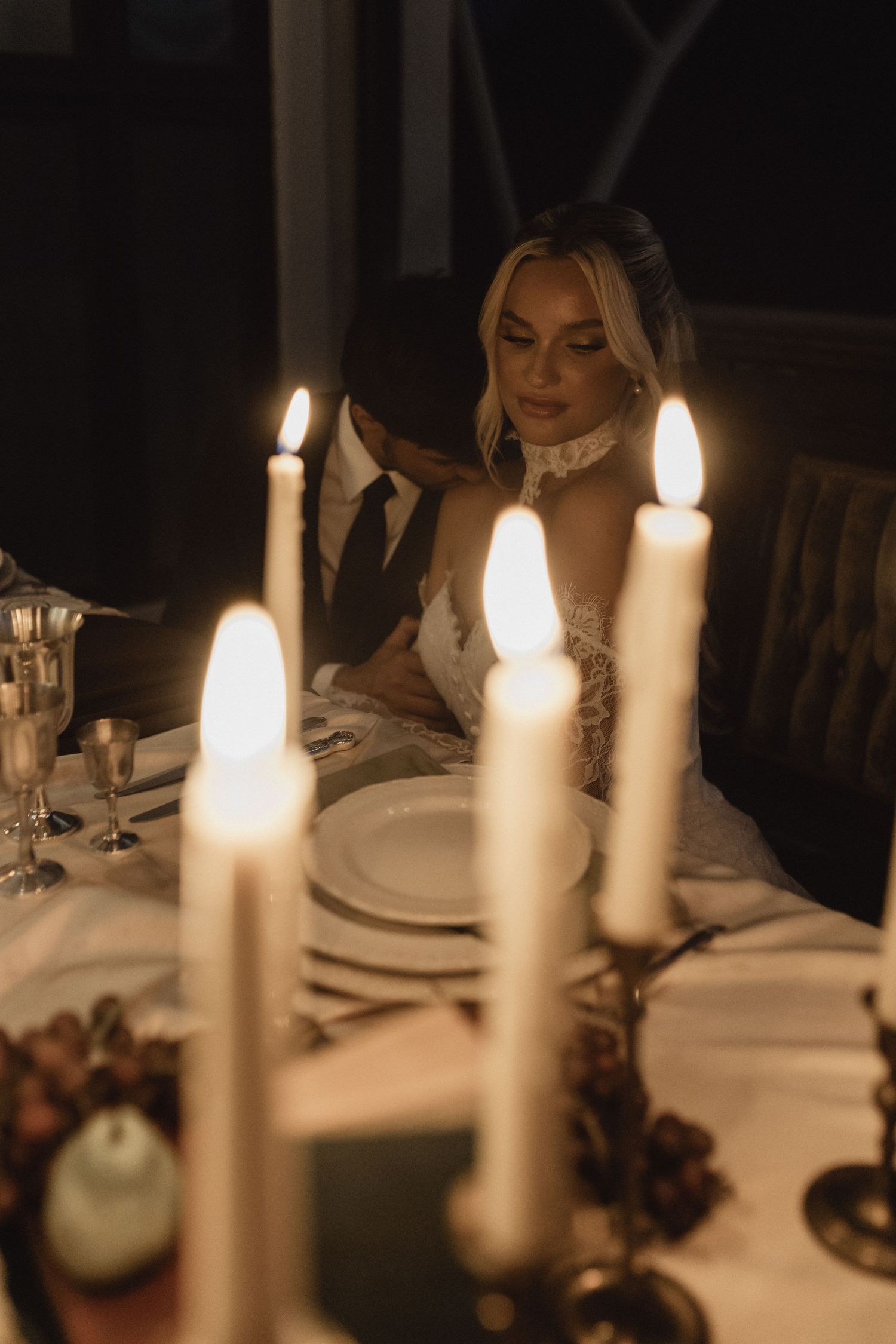 Bride and groom share an intimate candlelit dinner, with tall white candles glowing warmly at an elegantly set table.