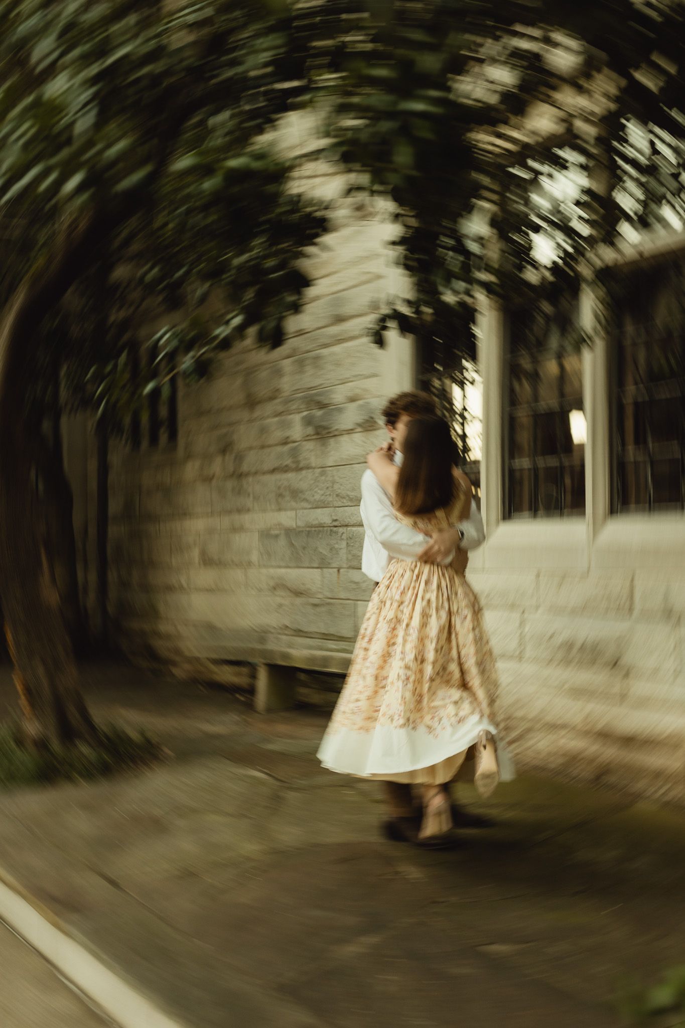 A woman in a floral midi skirt twirls under a lush green vine tunnel beside a rustic white building.