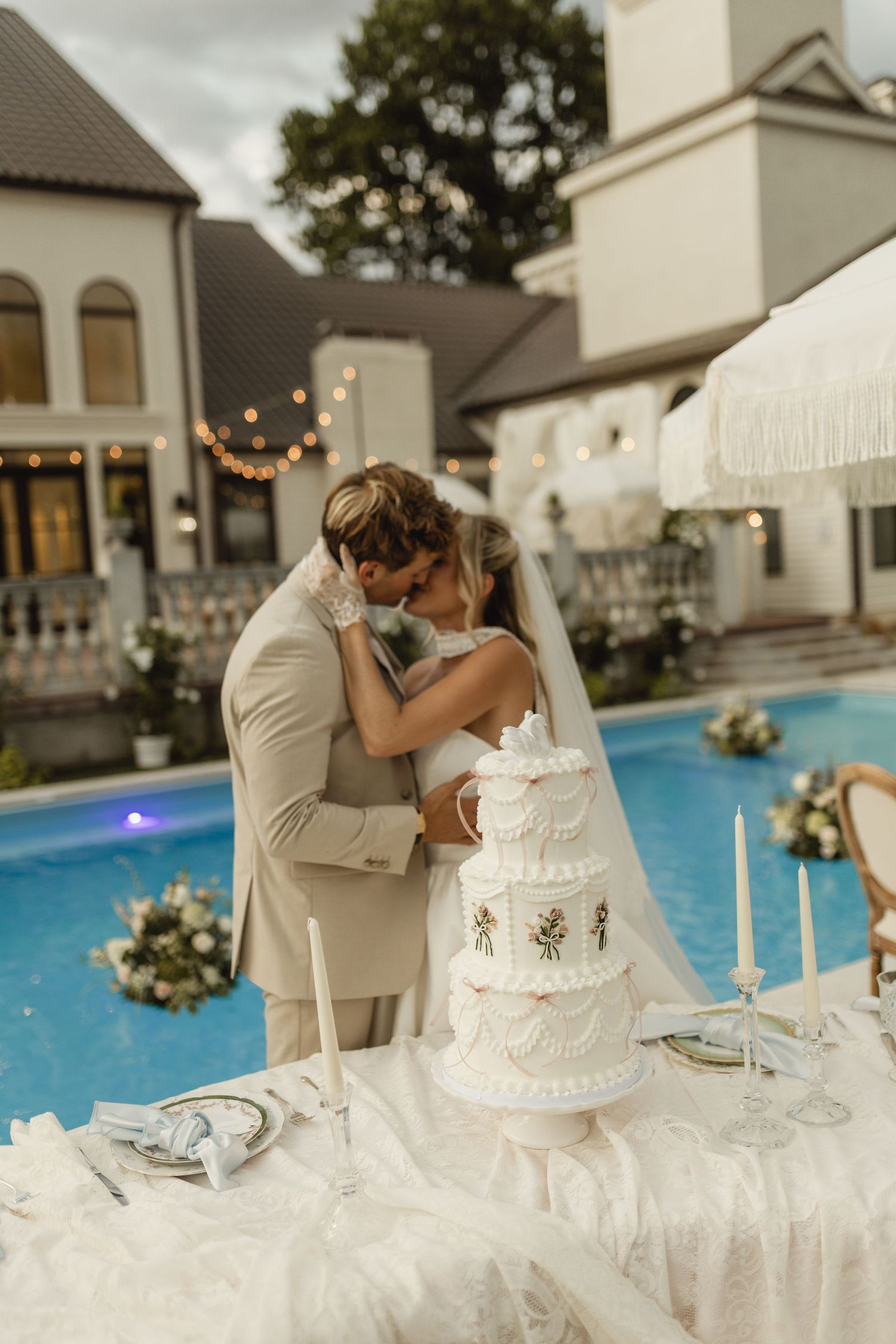 Bride and groom kissing beside a tiered white wedding cake near a pool at an elegant outdoor venue.
