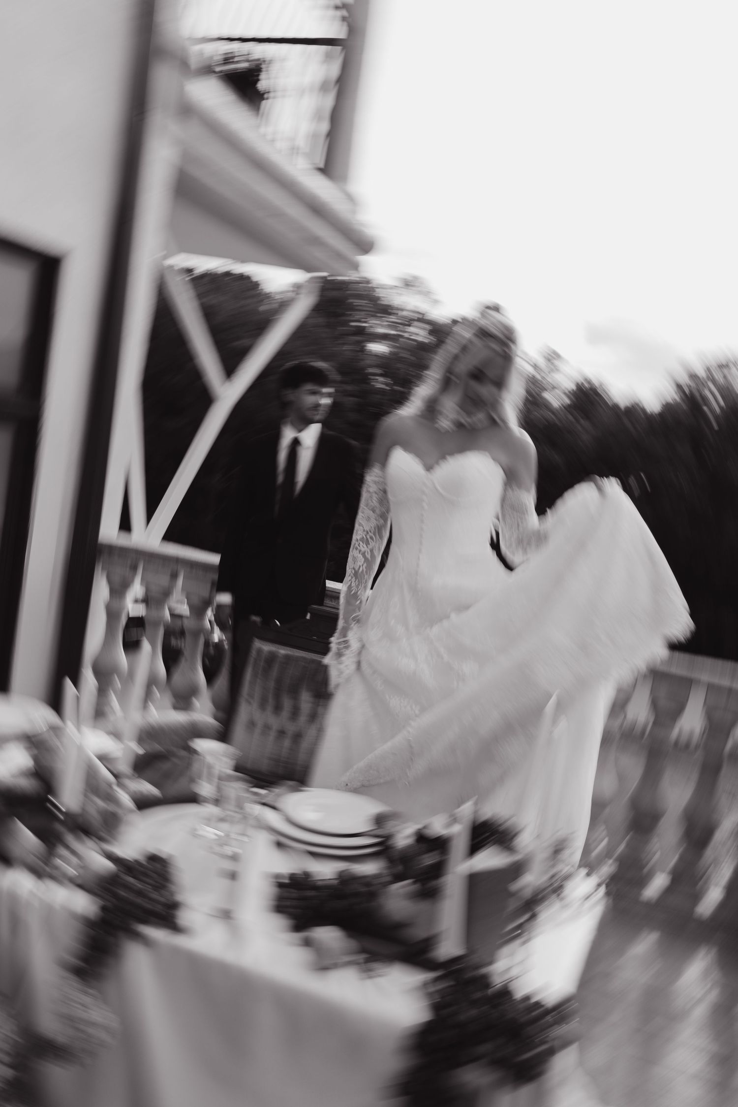 Black and white photo of a bride in a flowing gown walking with groom at an outdoor wedding reception table.
