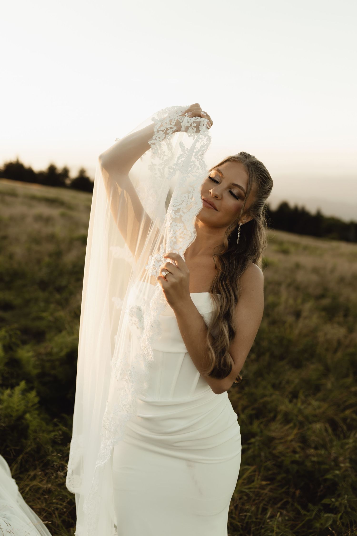 Bride in white gown holding lace veil at golden hour in a field.