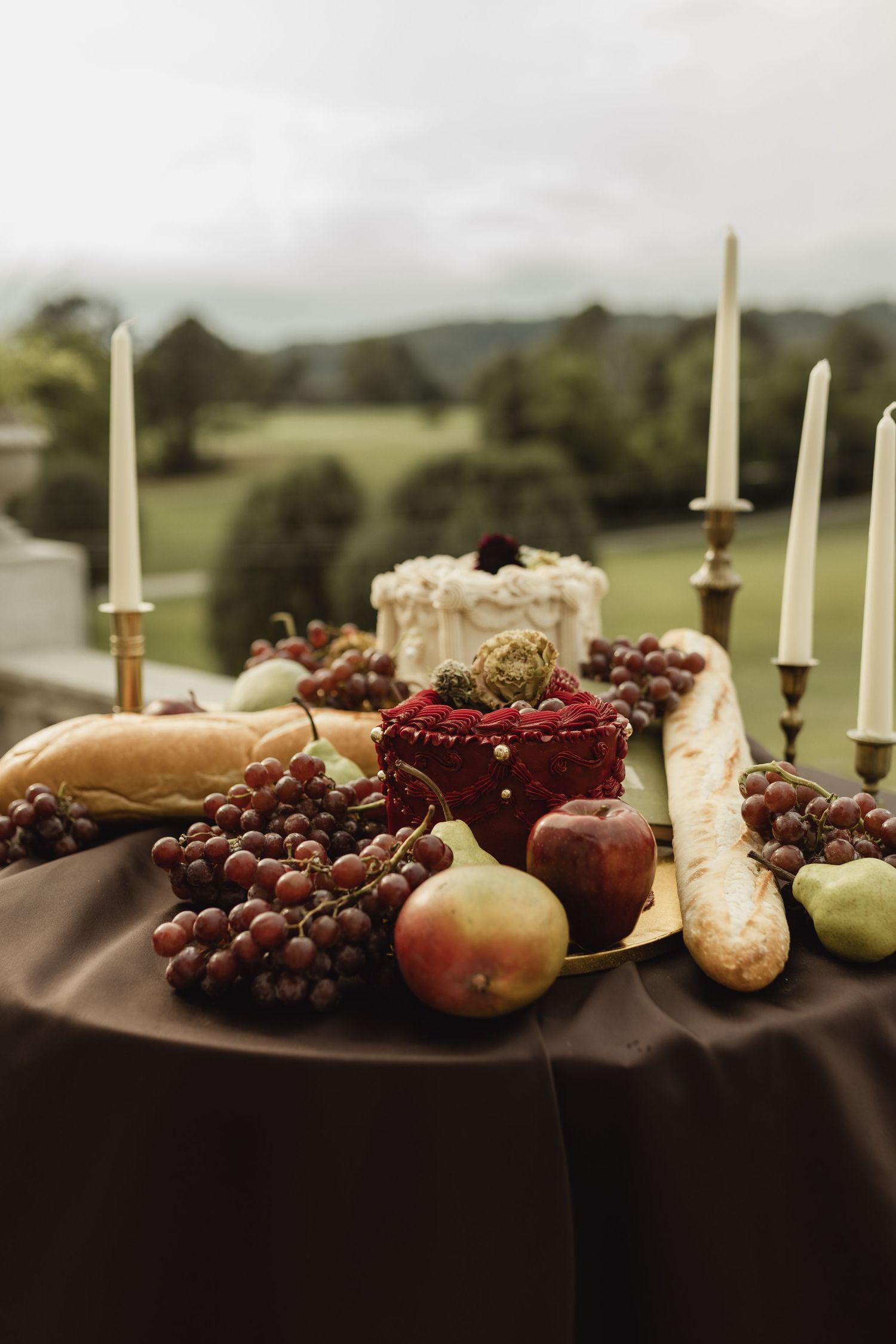 Elegant outdoor table display with grapes, pears, baguette, candles, and cake against a lush green landscape backdrop.