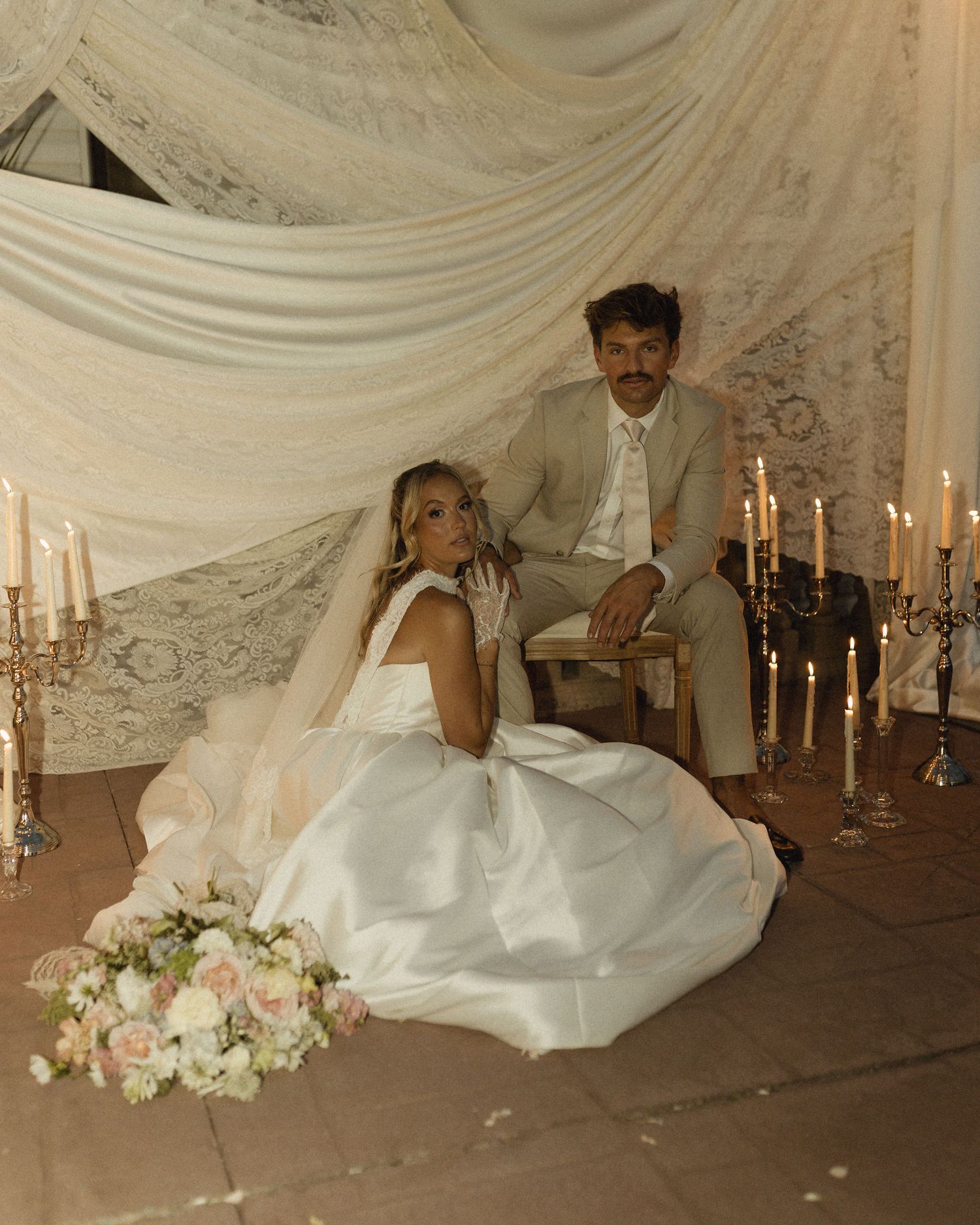 Bride in white gown sits on floor beside groom in tan suit, surrounded by candles and lace backdrop.