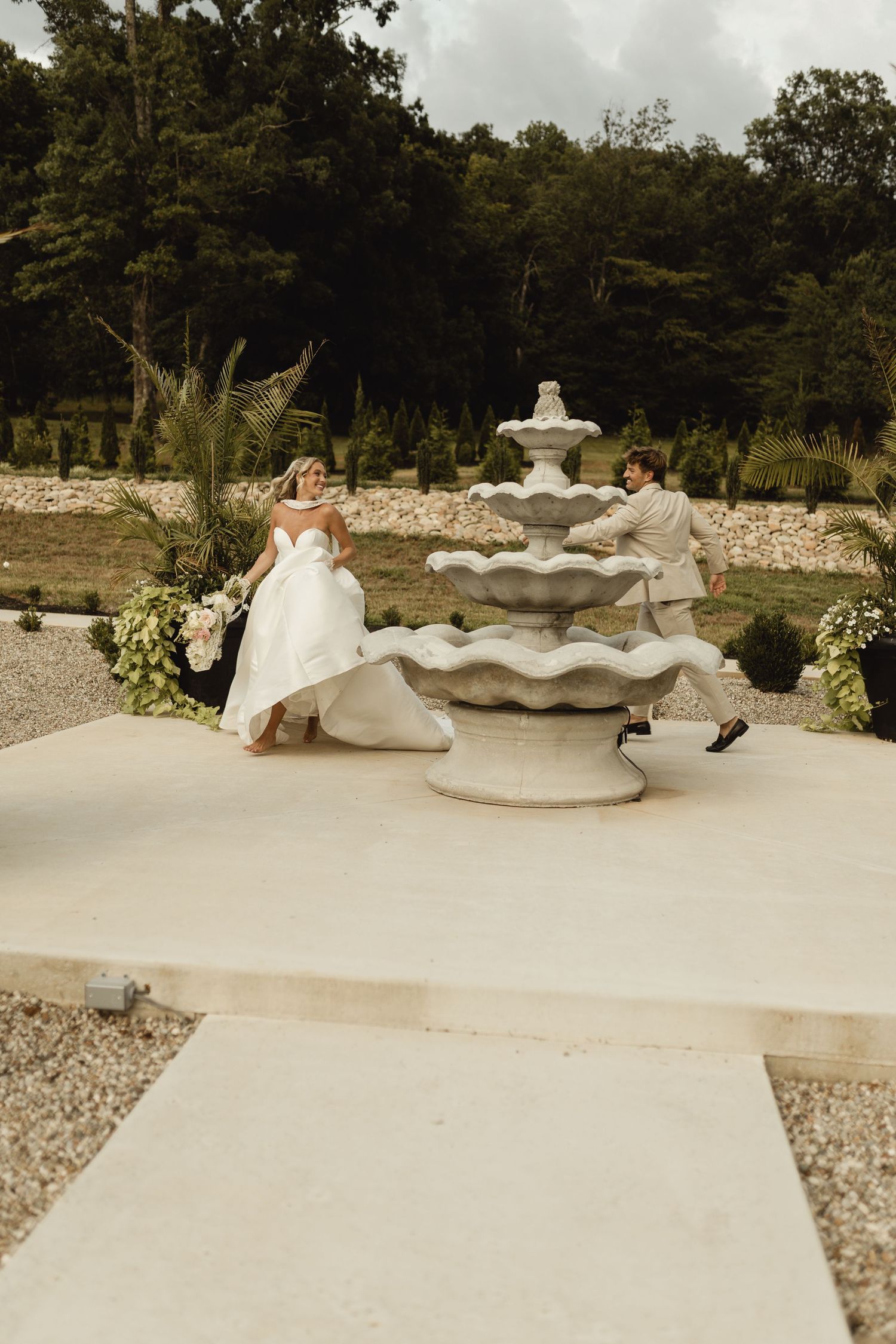 Bride and groom playfully chase each other around a tiered stone fountain at an outdoor wedding venue.